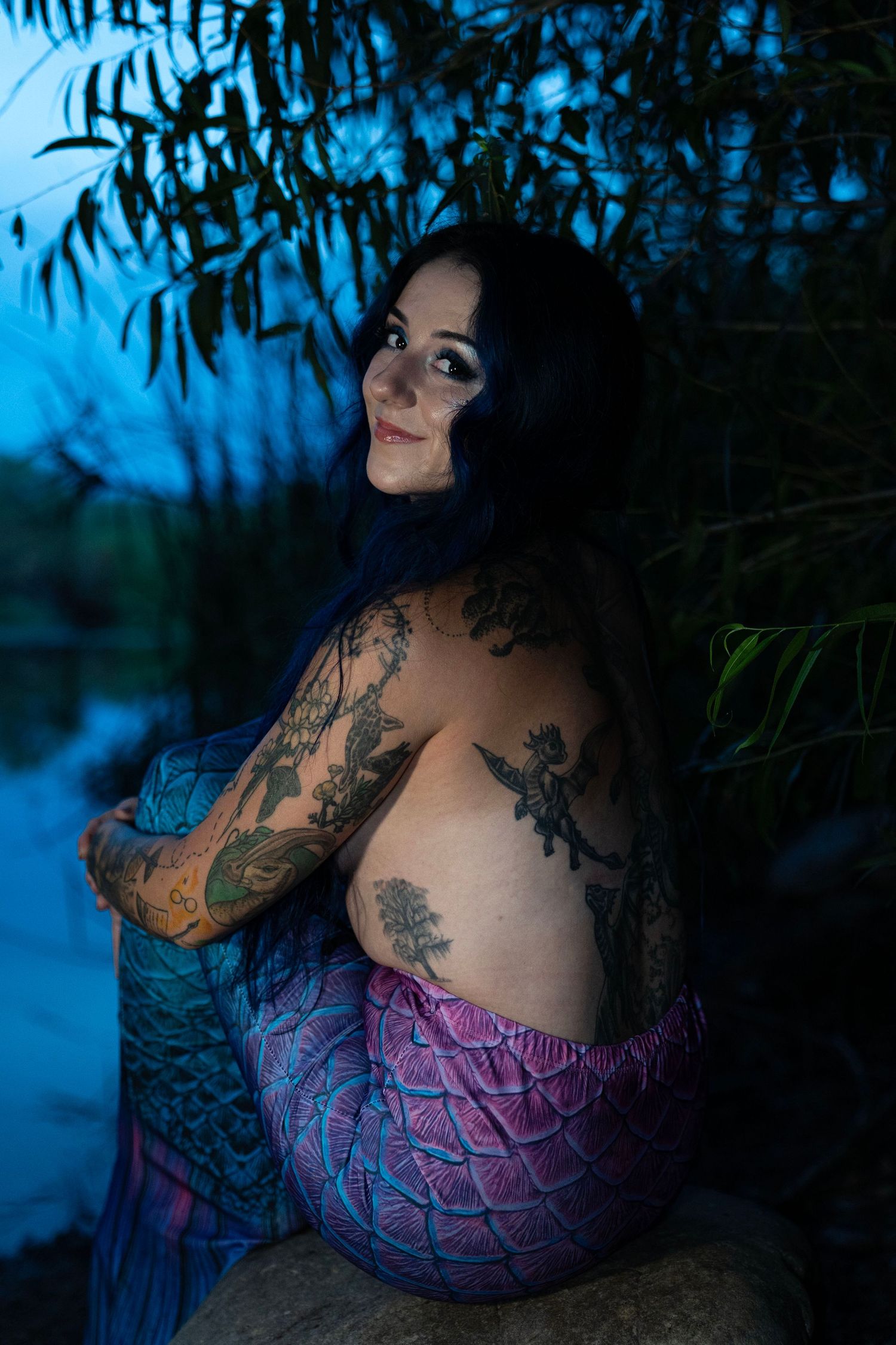 Heavily tattooed woman in pink and blue mermaid tail posing by a river in blue twilight.