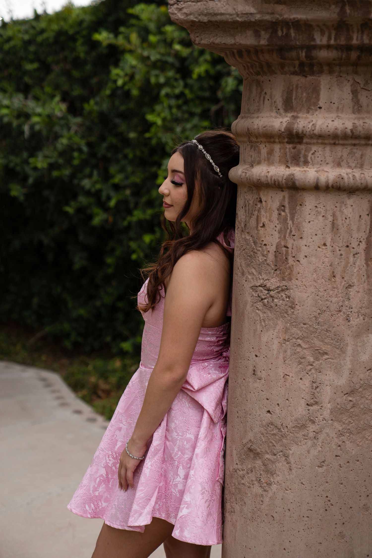 Young woman in pink dress leaning against stone column in garden, wearing crystal headband, side profile view.