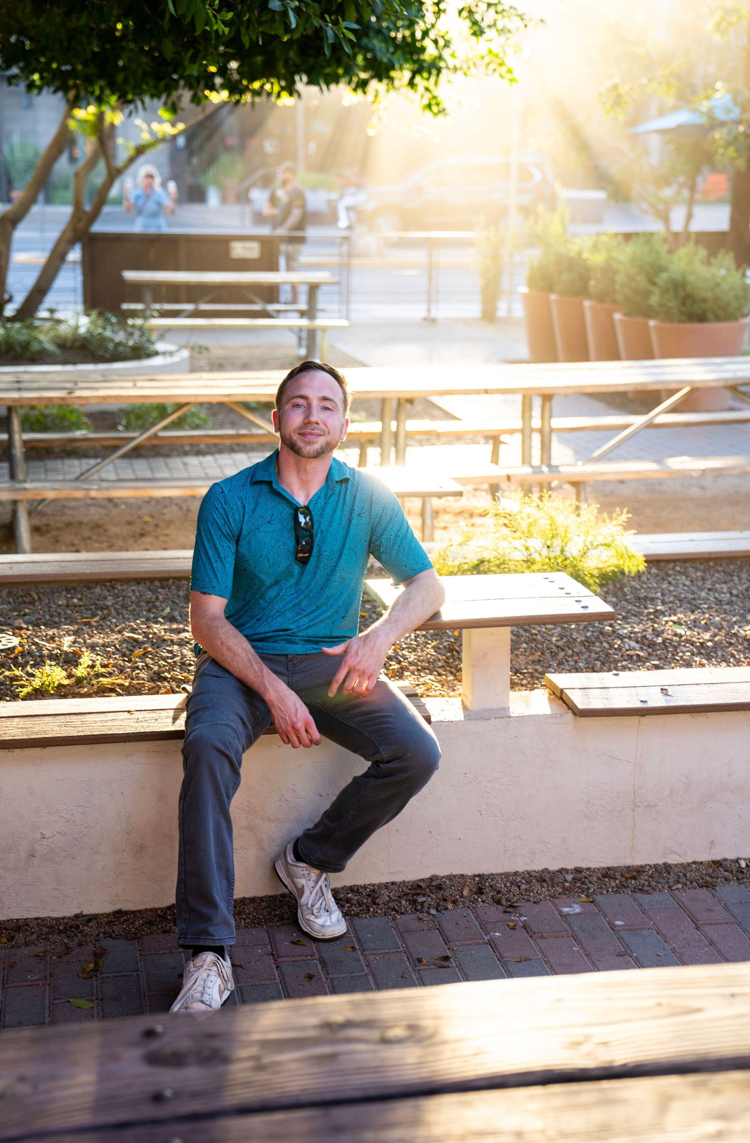 Man in teal polo shirt sitting on outdoor bench wall smiling in sunny plaza setting.