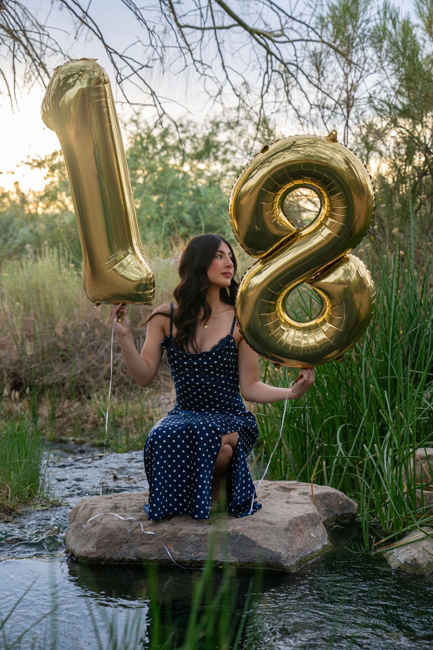 Woman in polka dot dress holding gold number 18 balloons by stream.