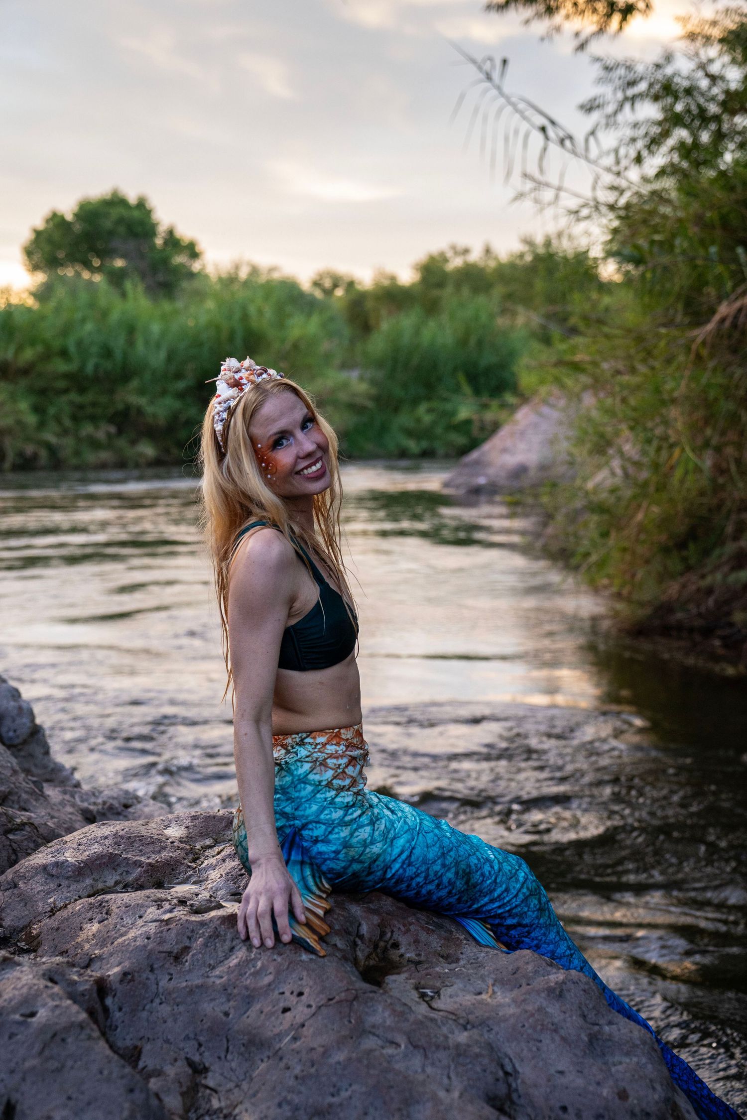 A smiling blonde woman in a blue mermaid tail and floral crown sits on rocks by a sunset river. Mermaid photoshoot, fantasy photoshoot.