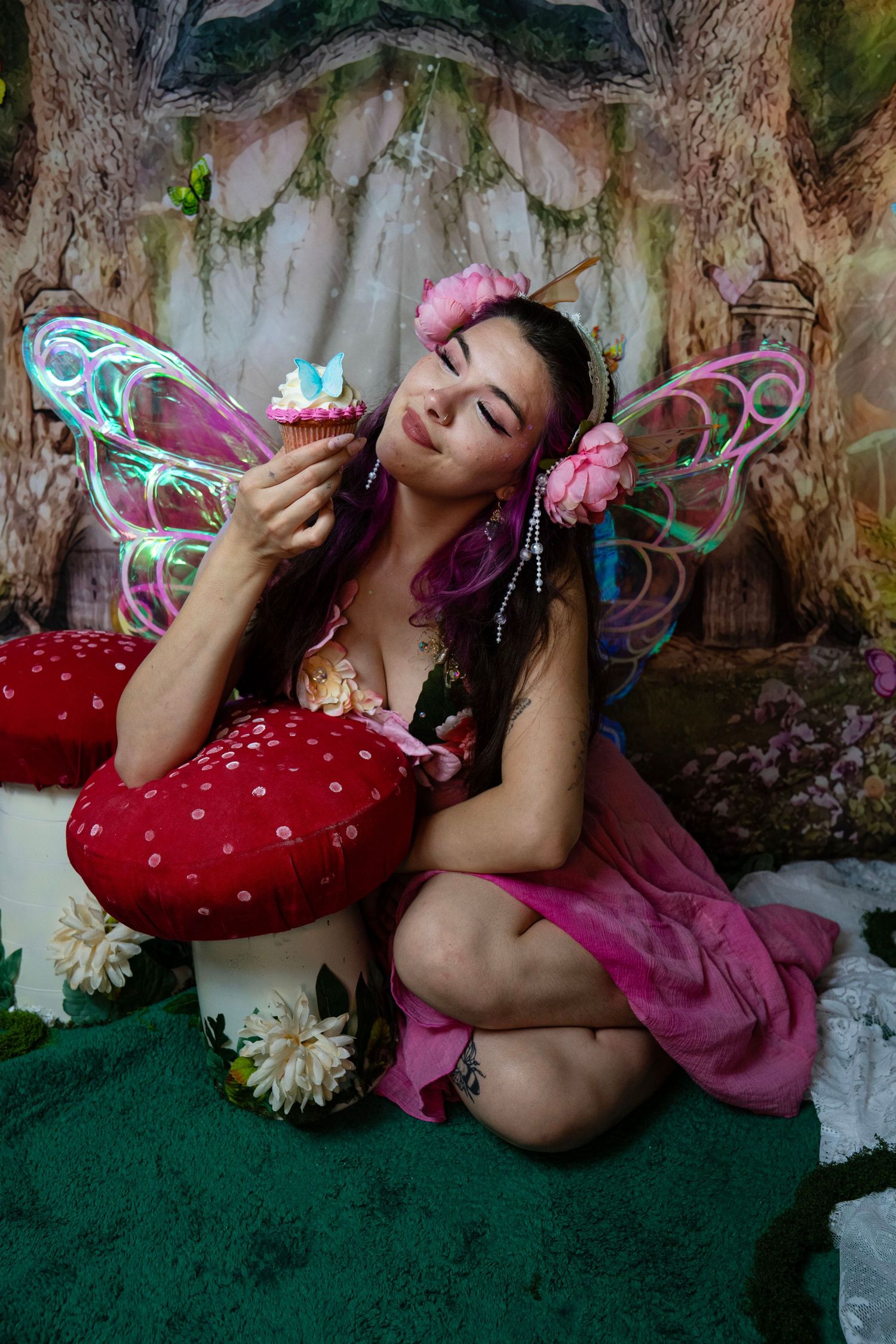 Fairy with glowing wings and pink floral dress holds a cupcake with eyes closed in a dreamy enchanted forest setting. Fantasy Photoshoot