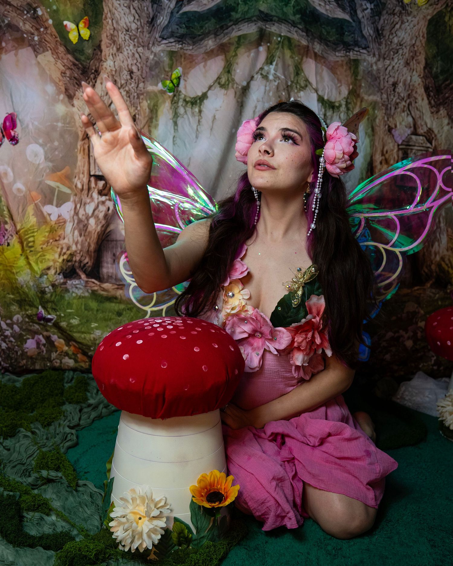 Woman dressed as fairy with colorful wings reaches upward while kneeling among mushrooms in magical woodland scene. Fantasy Photoshoot