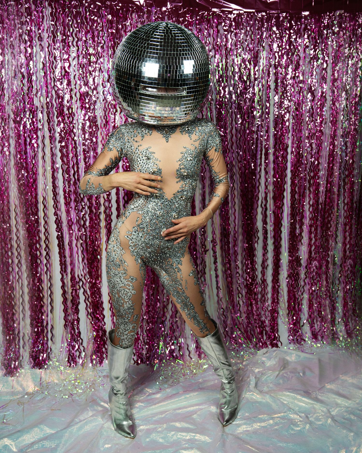 Model in silver crystal jumpsuit and boots poses with large disco ball covering head.