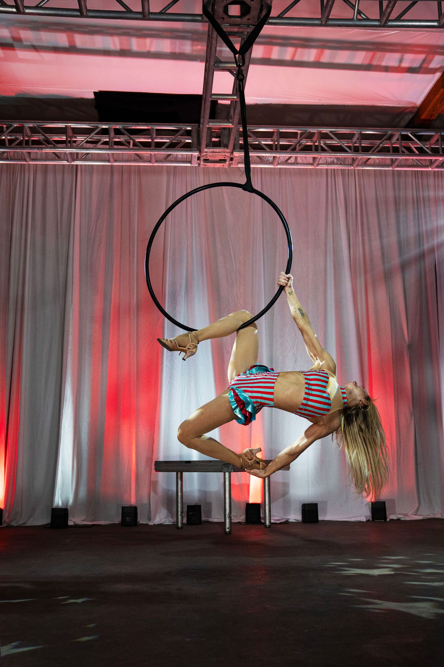 Flexible female aerialist performing a backbend on a suspended lyra hoop under red stage lighting. Circus Photoshoot