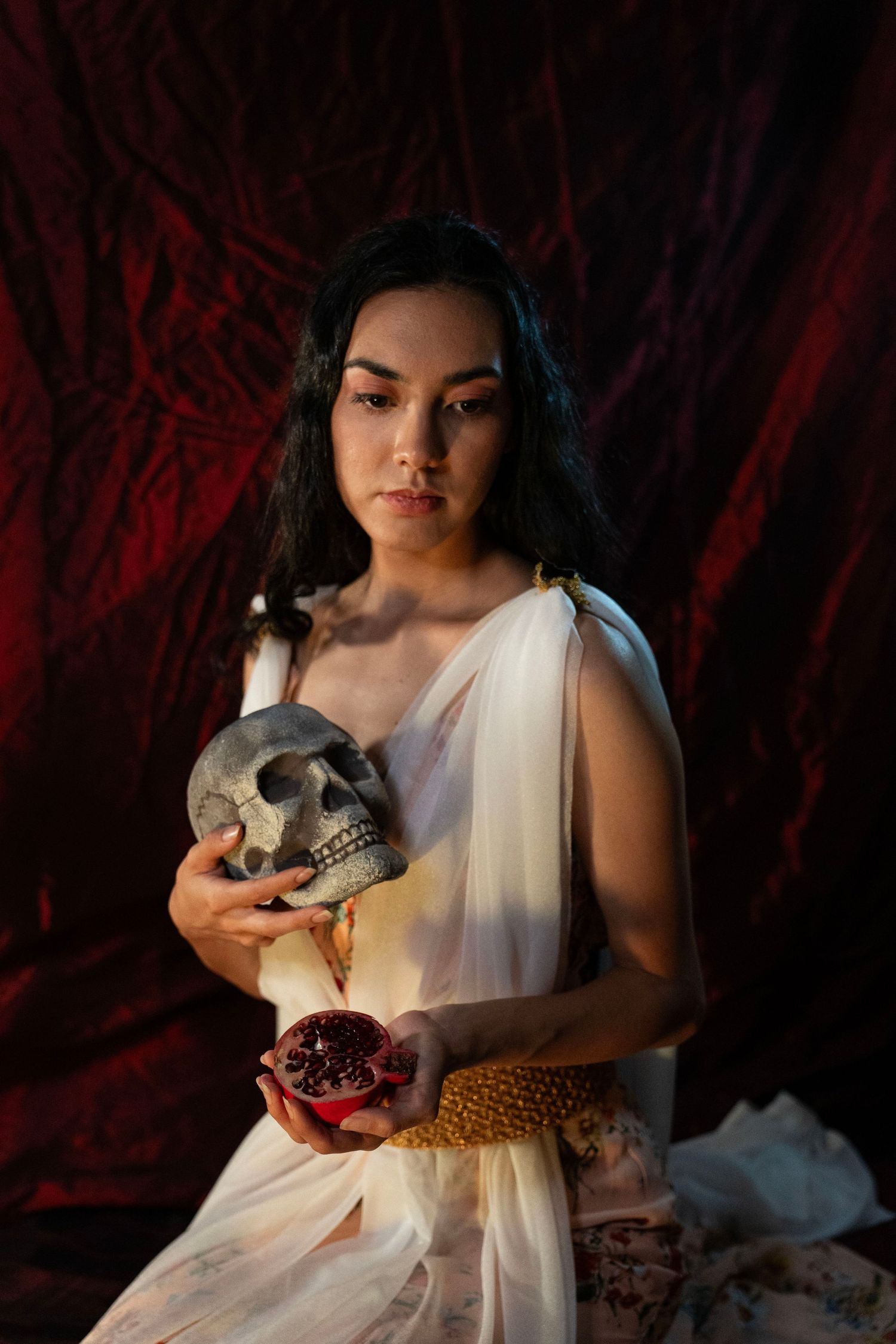 Dark-haired woman in a white draped gown holds a stone skull and a pomegranate half in a moody, dramatic red velvet setting.