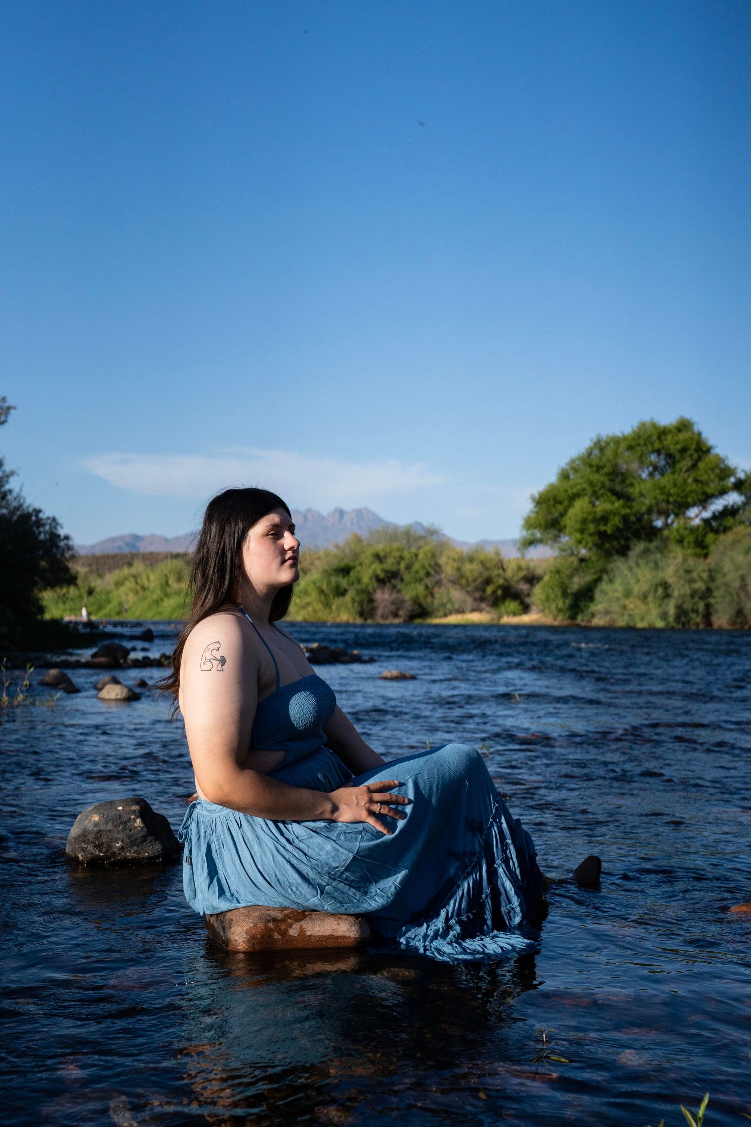 Woman in blue dress sitting on river rocks with mountain backdrop and clear blue sky.