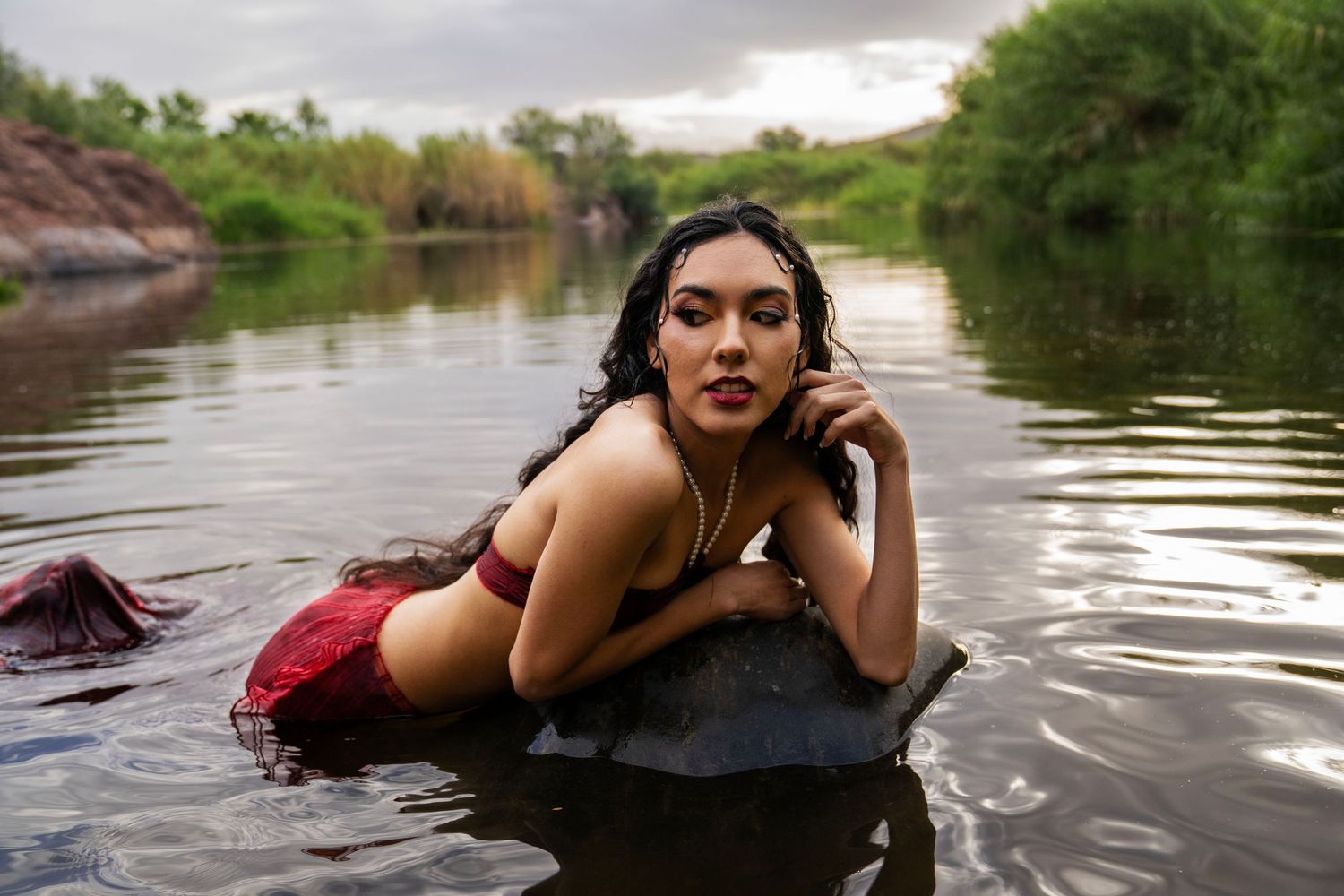 A young woman with wet hair leans on a rock in a river, wearing a red outfit in an outdoor mermaid-themed photo session.