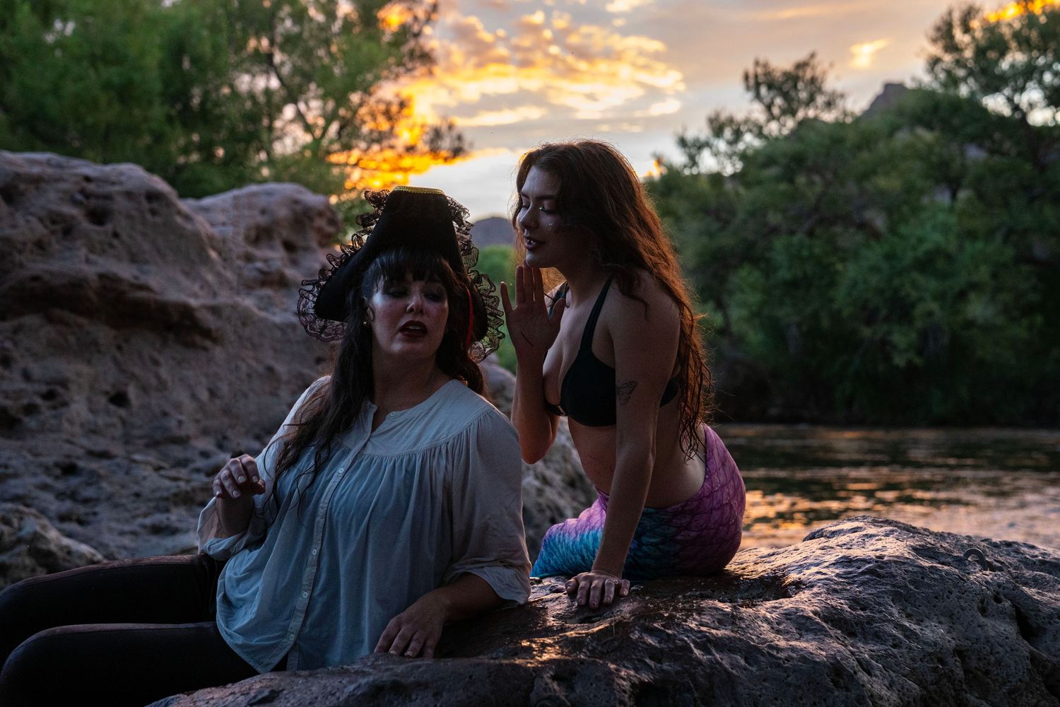 Two women in pirate and mermaid costumes sit on rocks by a river during a colorful sunset. Mermaid photoshoot, fantasy photoshoot.
