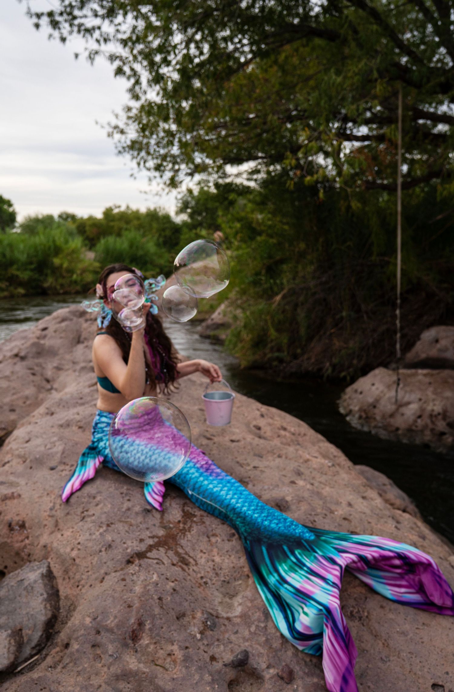 Woman in mermaid costume blowing giant soap bubbles while reclining on rocks beside a flowing river. Fantasy Photoshoot