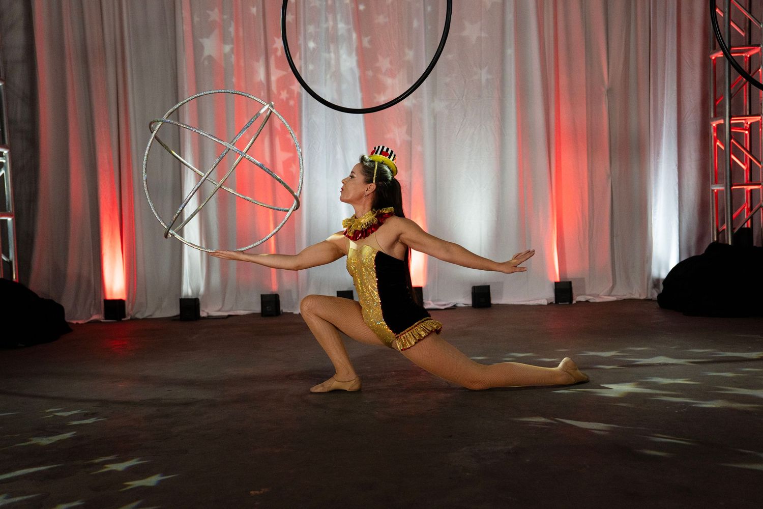 Circus performer in gold costume doing splits pose beside aerial hoop on stage.