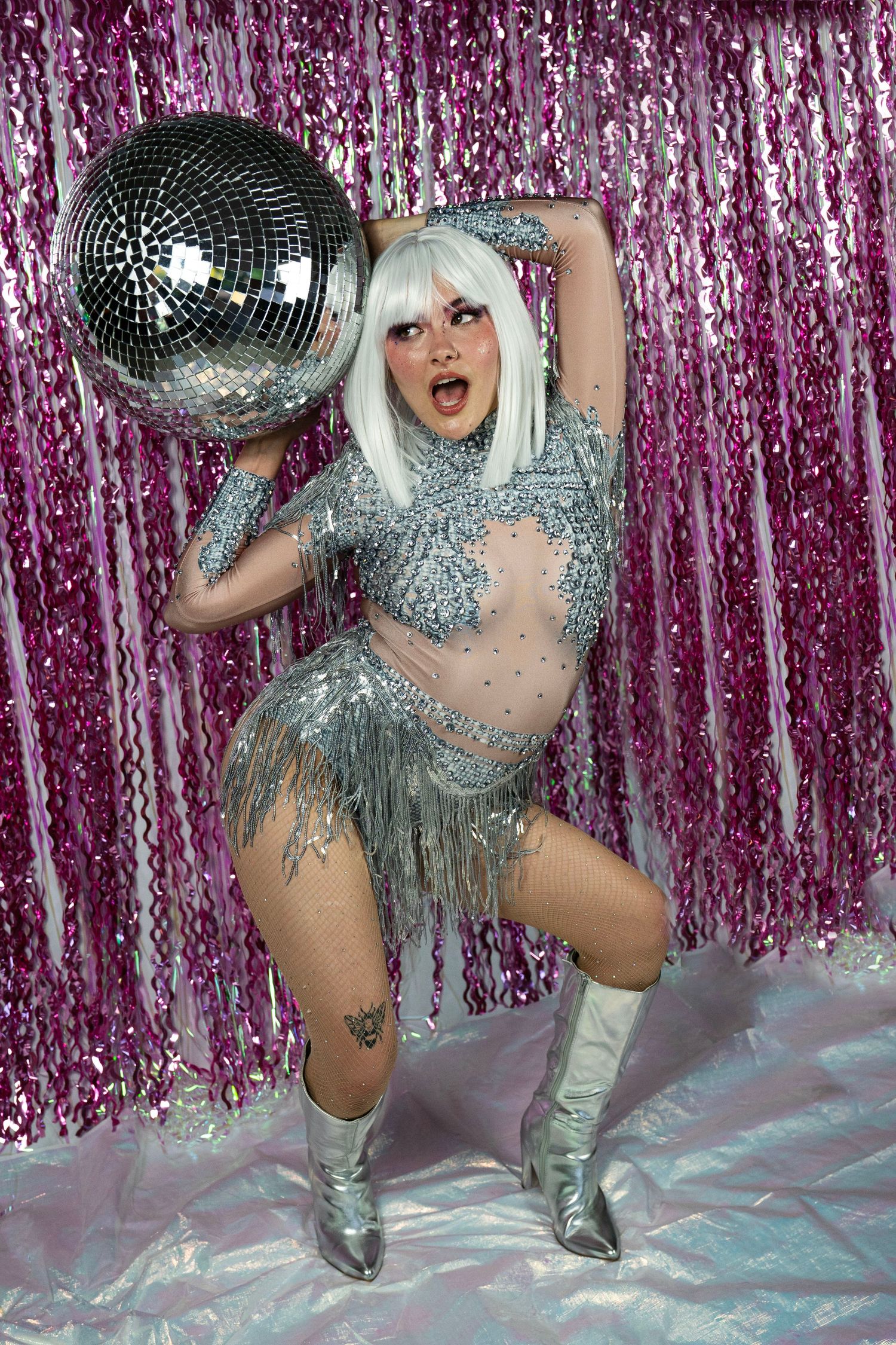 Female performer in silver bedazzled fringe bodysuit crouches holding large mirror disco ball against pink tinsel curtain.