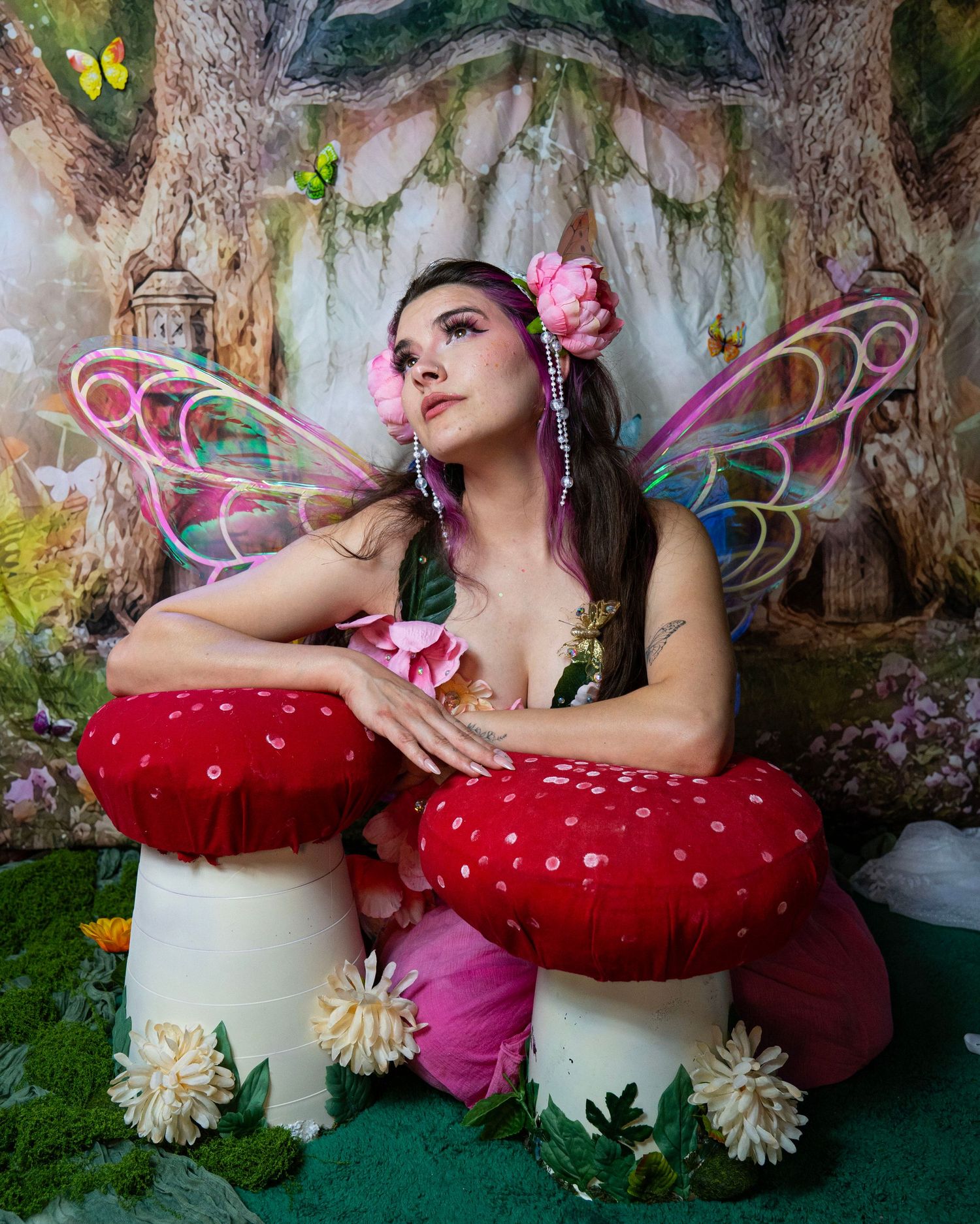Fairy cosplay model with colorful wings and pink hair accessories leans on giant red mushrooms in fantasy setting. Fantasy Photoshoot