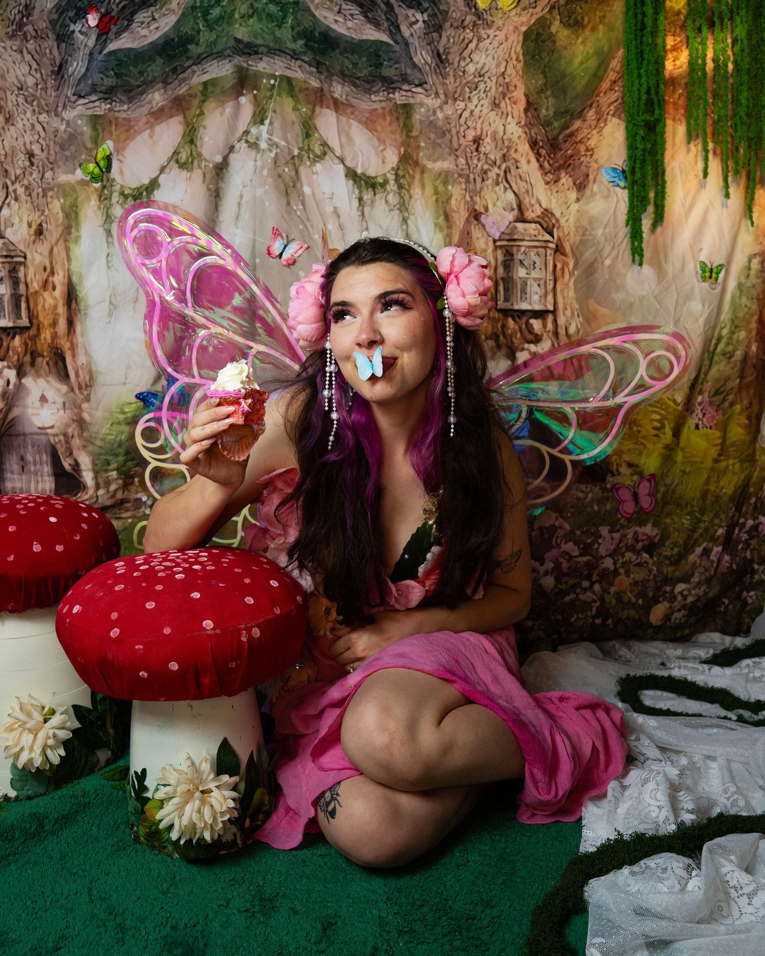 Fairy costumed woman with pink wings holds cupcake gazing upward with blue butterfly on lips in forest set. Fantasy Photoshoot