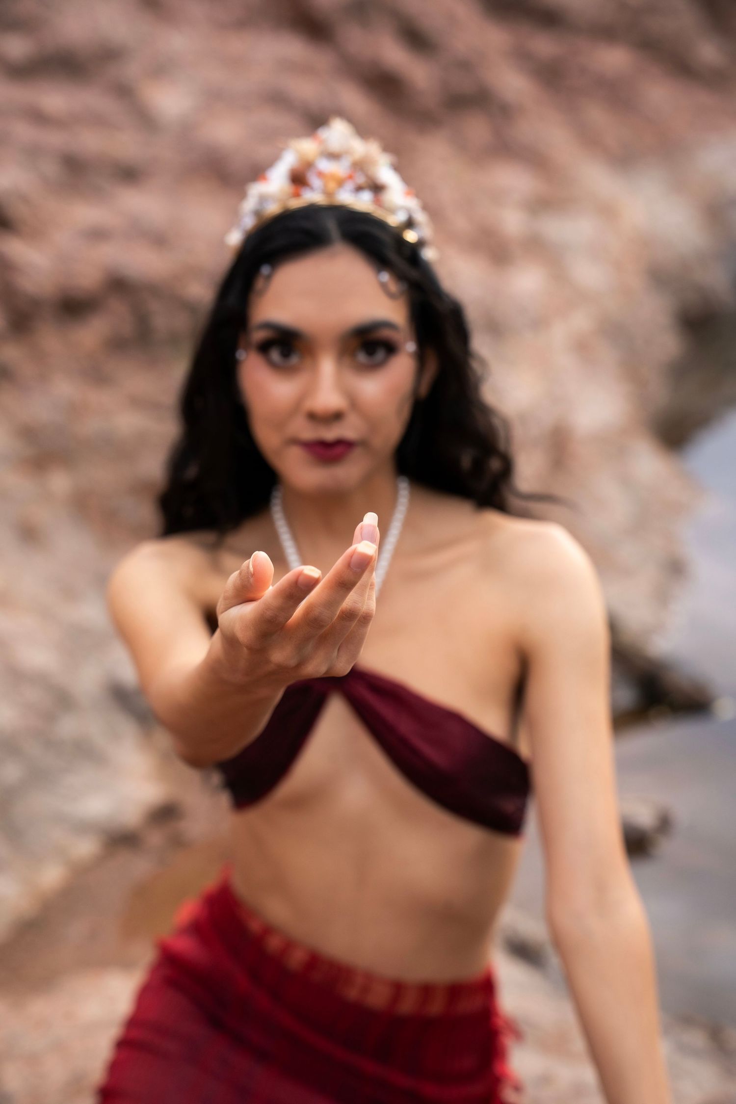 A woman in a red mermaid costume and shell crown extends her hand toward the camera near rocky terrain.