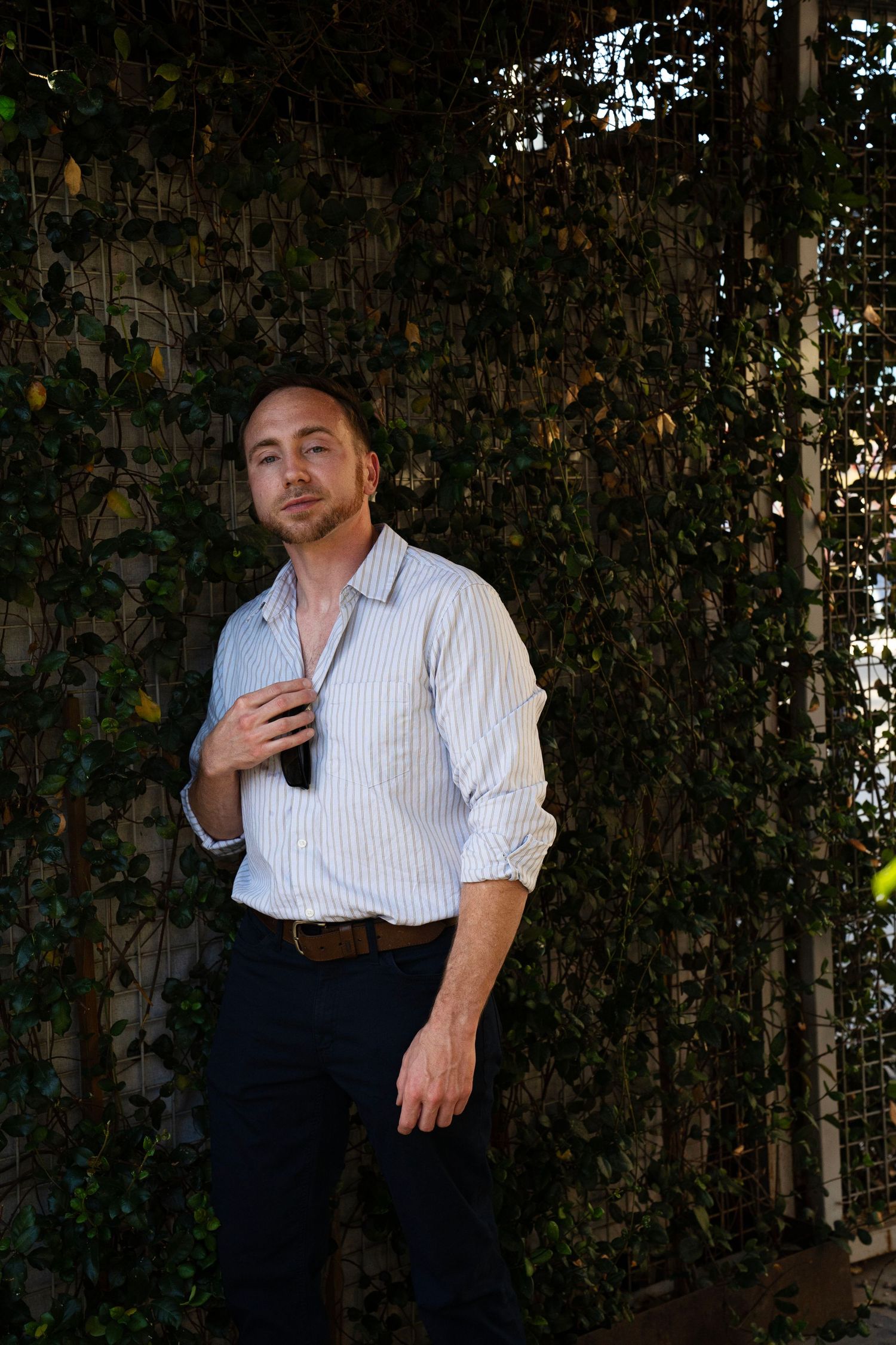 Man in striped shirt standing against ivy-covered wall outdoors, holding sunglasses in a relaxed pose.
