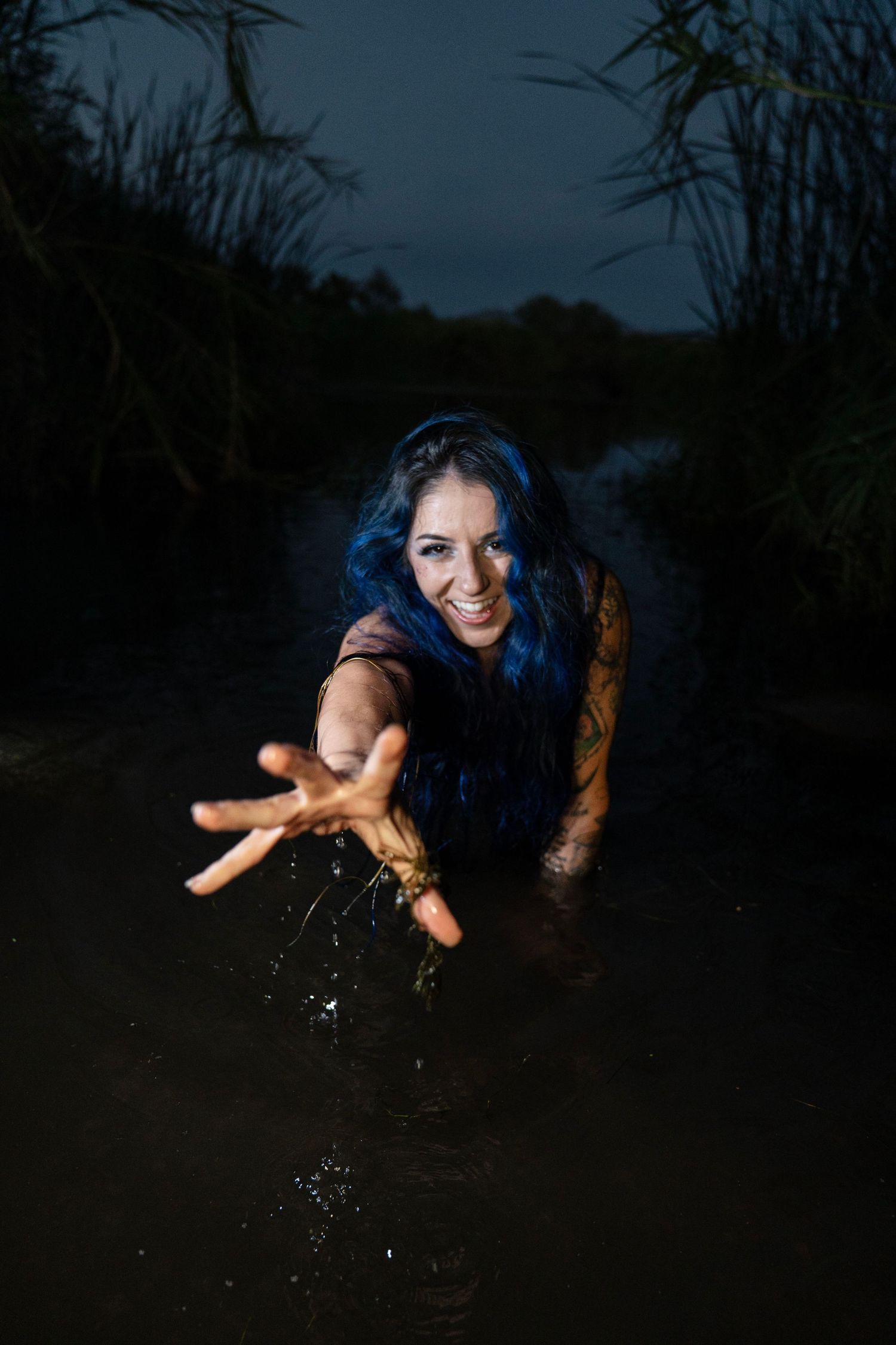 Smiling woman with blue hair reaches toward the camera while standing in dark water at dusk, surrounded by reeds.