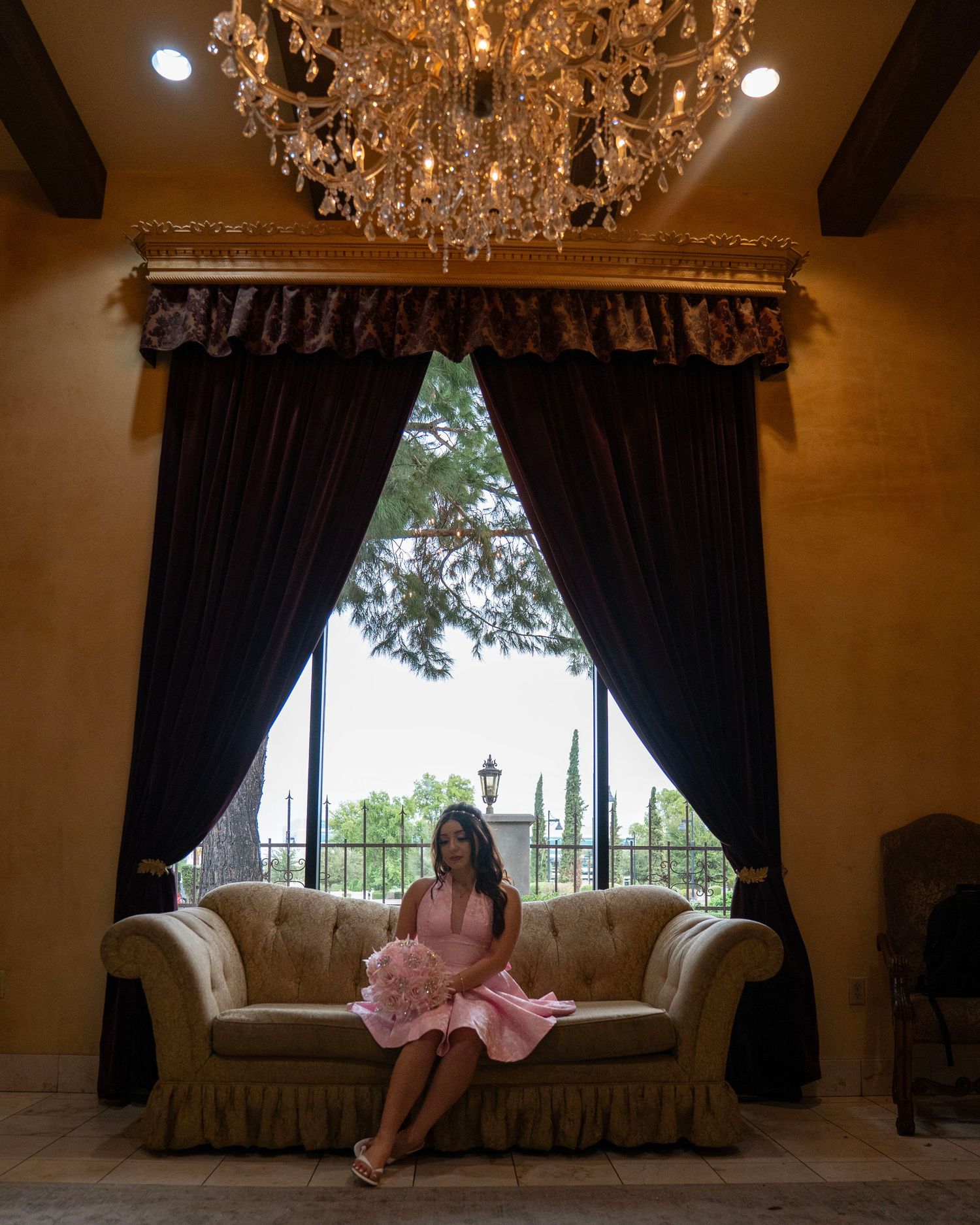 Woman in pink dress sitting on a loveseat beneath an ornate crystal chandelier by a draped window.