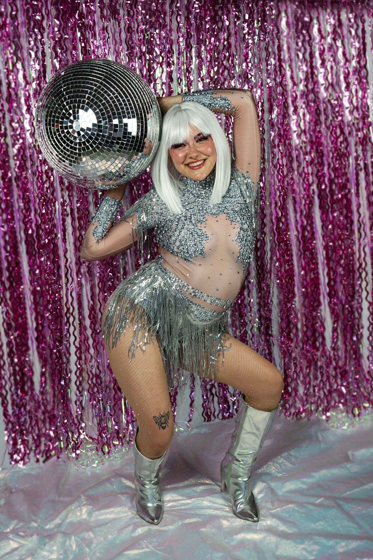 Female performer in silver sequin fringe bodysuit and white wig poses with large disco mirror ball and silver boots.