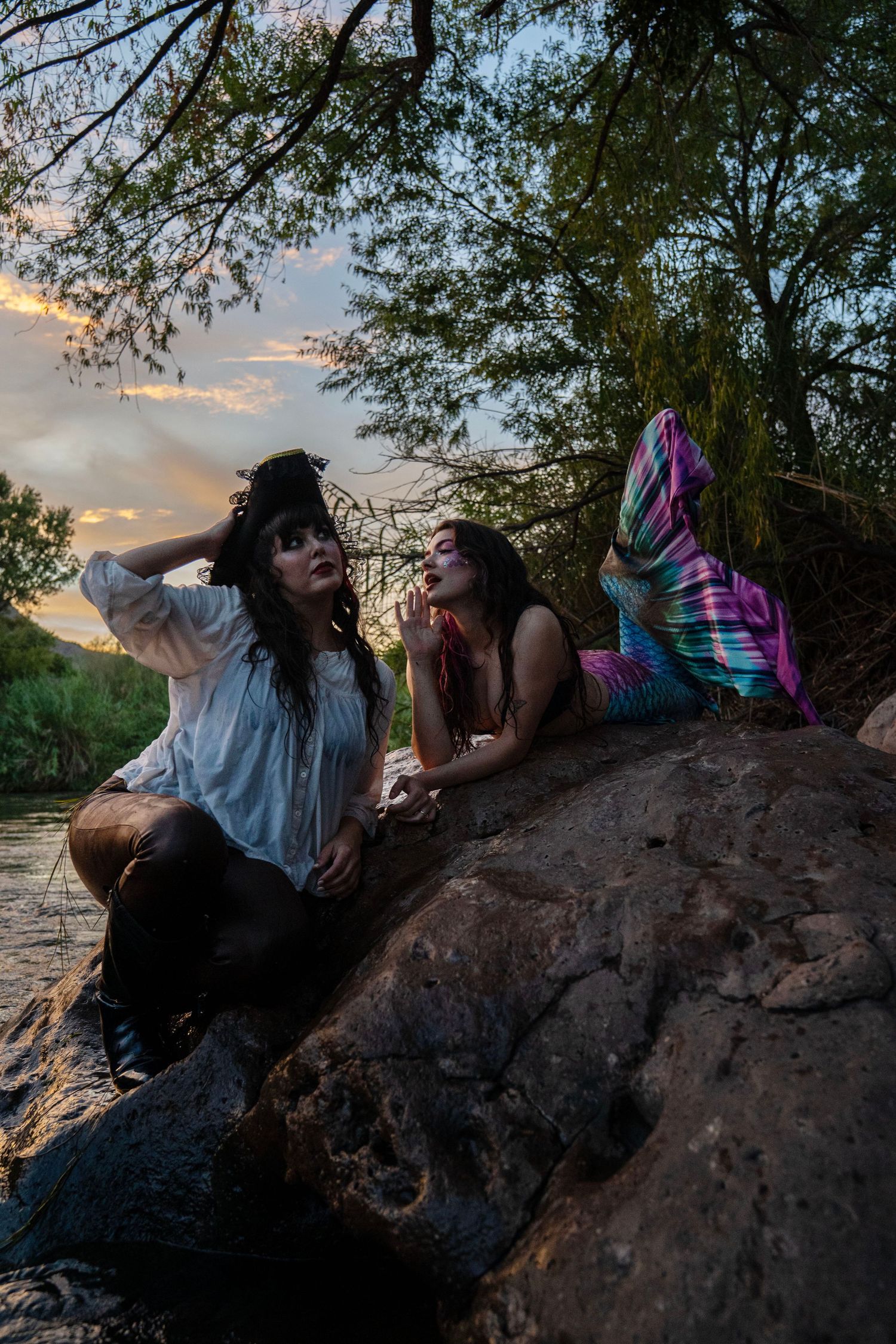 A pirate and mermaid costume duo pose on riverside rocks at sunset with dramatic cloudy sky. Mermaid photoshoot, fantasy photoshoot.