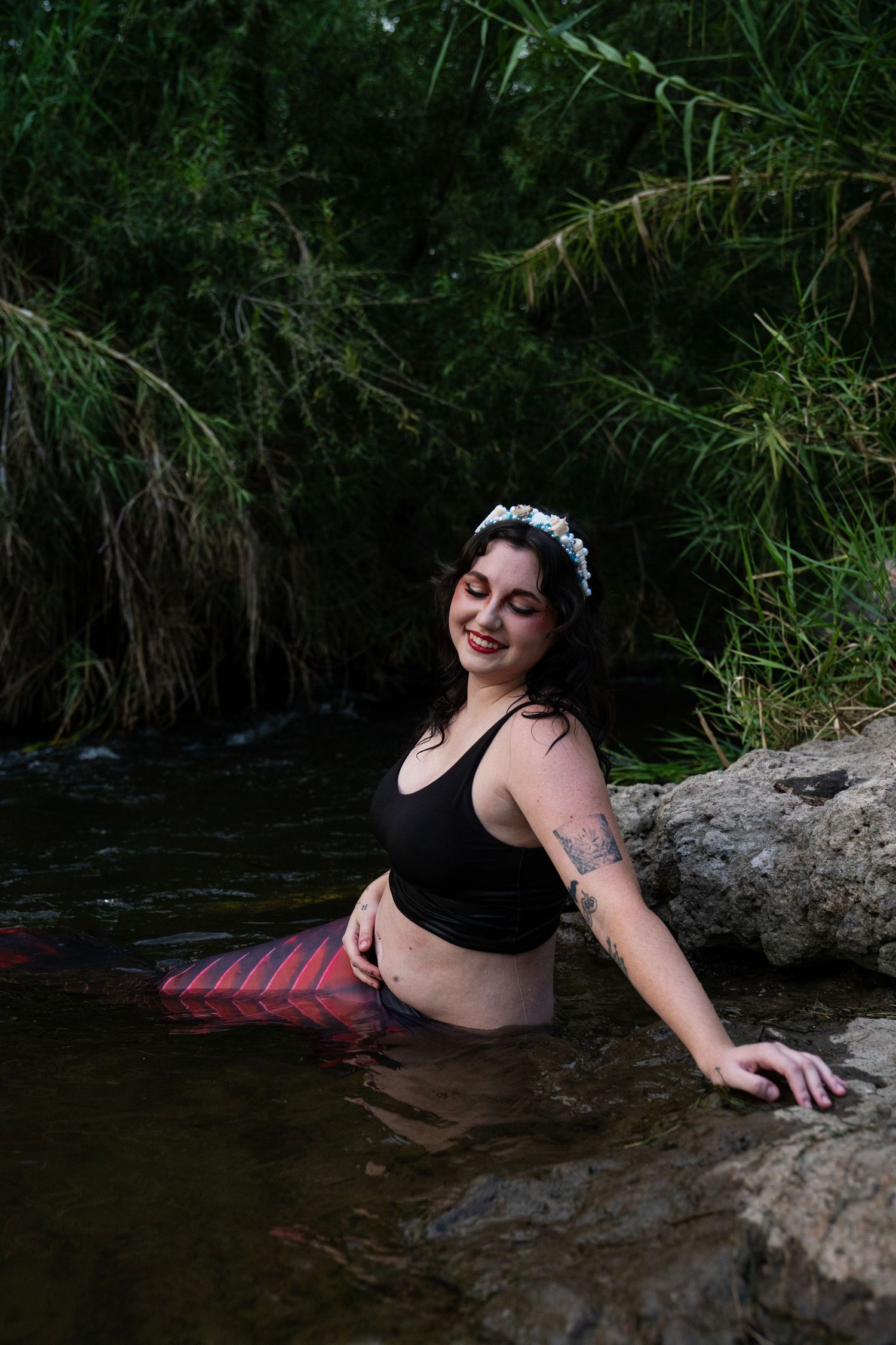 Smiling woman in black bikini top with red mermaid tail and floral crown in river. Fantasy Photoshoot