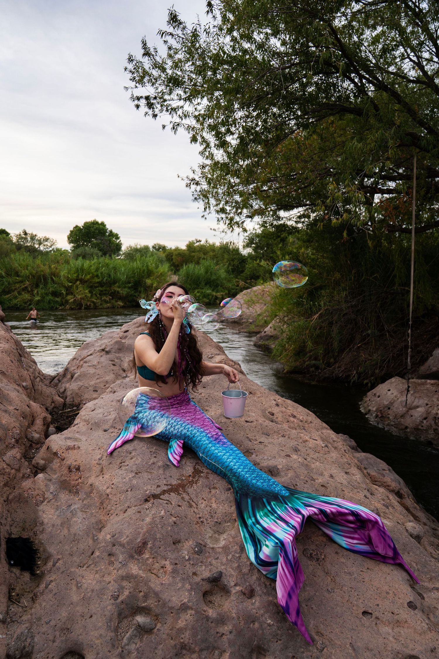 Mermaid cosplay model in pink and blue tail blowing large soap bubbles on rocky riverbank outdoors. Fantasy Photoshoot