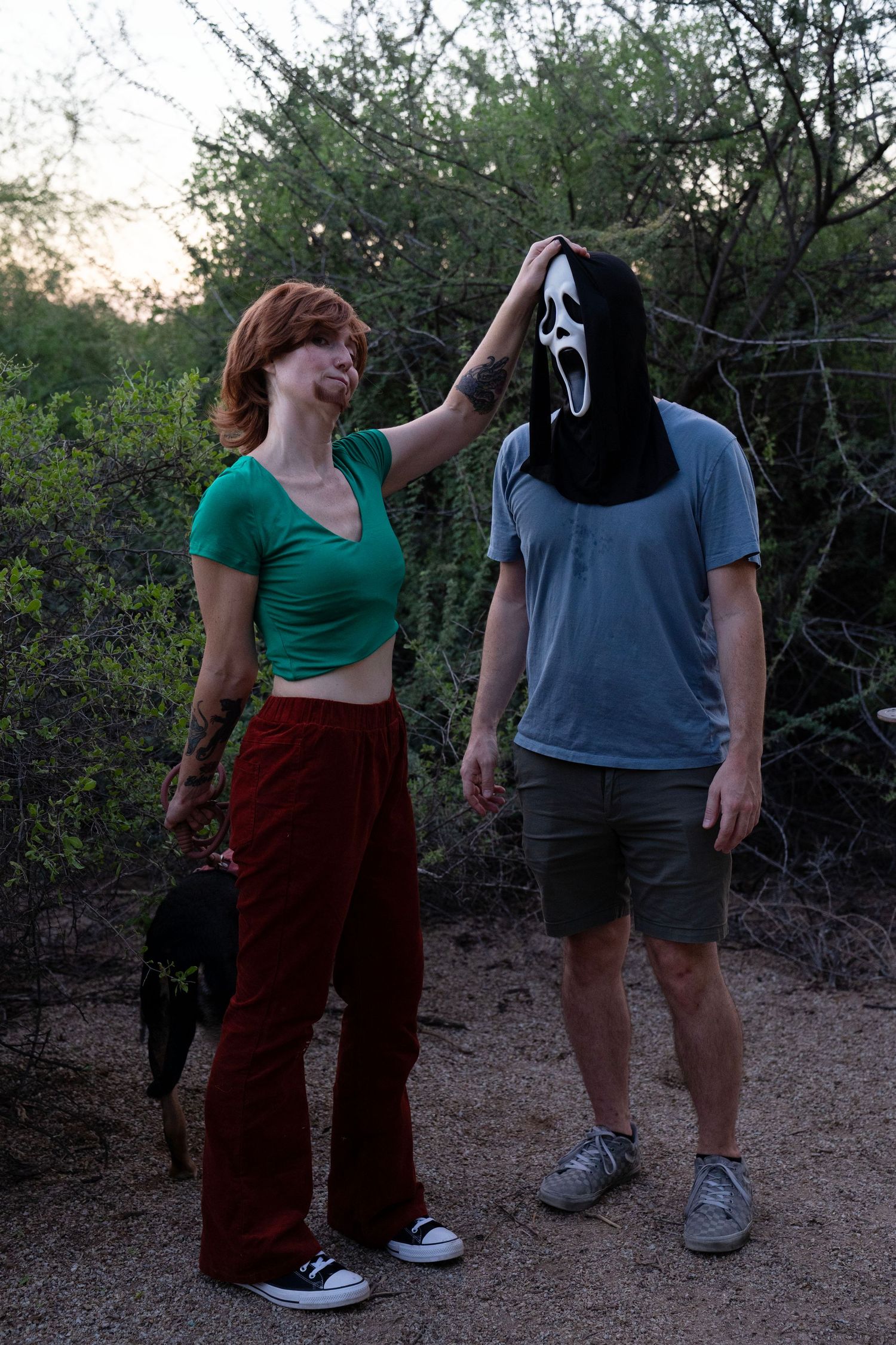 Woman in a green crop top and red pants rests her hand on a man wearing a ghost mask, standing outdoors near a dog among desert shrubs.