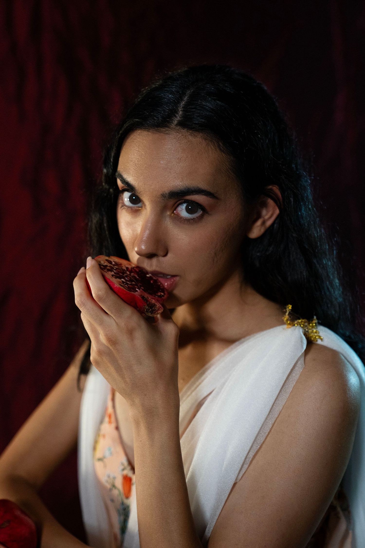 Woman with long dark hair in a white draped gown bites into a ripe pomegranate half against a deep red velvet backdrop.