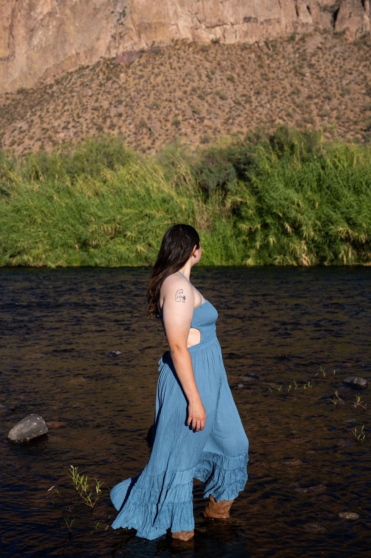 Woman in blue maxi dress wading in a river with lush greenery and rocky cliffs behind her.