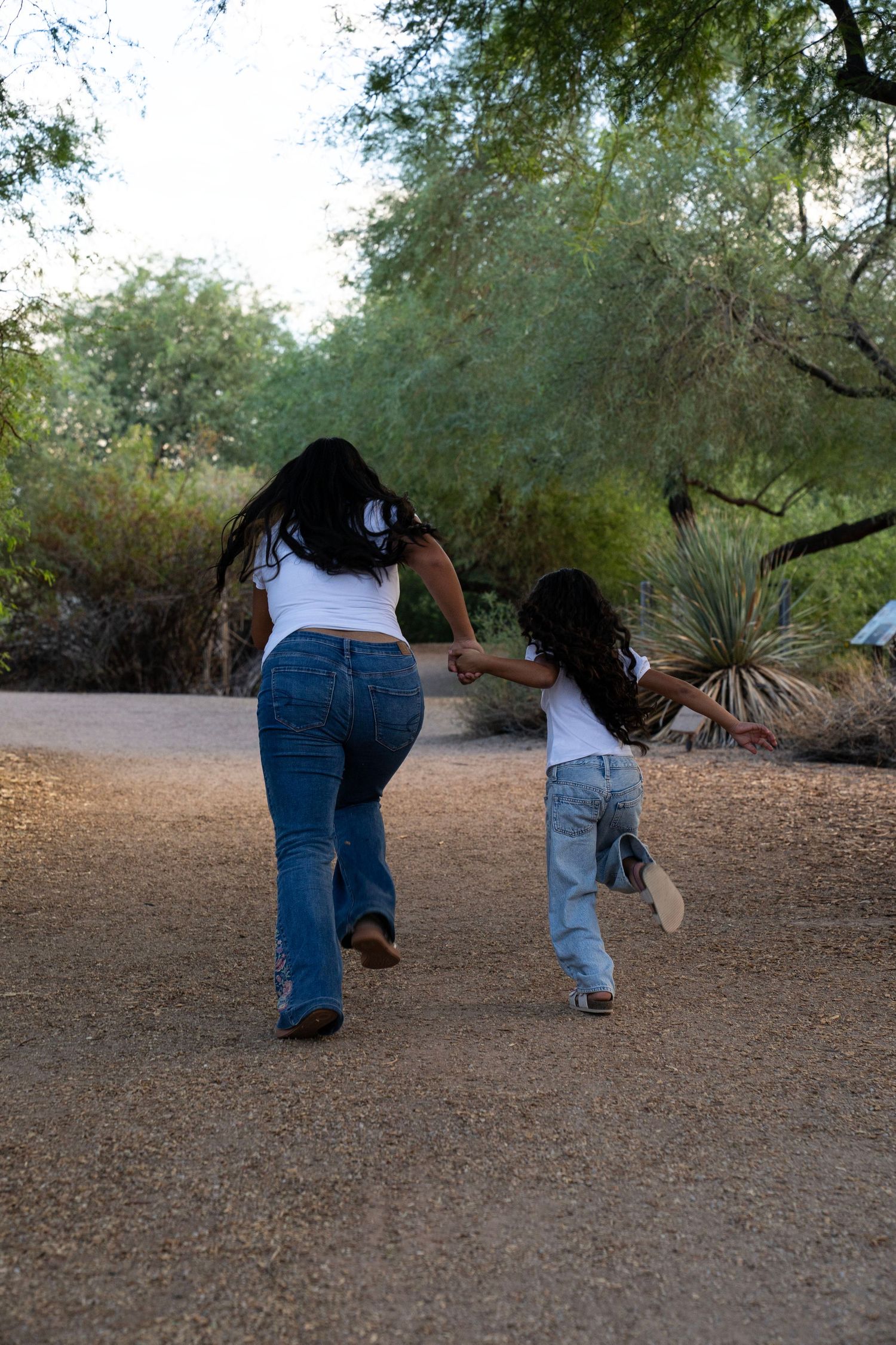 Mother and daughter in matching white tops and jeans walking hand in hand on a desert park path.