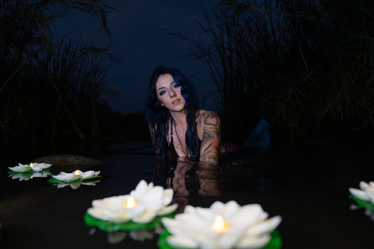 Tattooed woman in dark water gazing forward with white water lilies floating in the foreground.