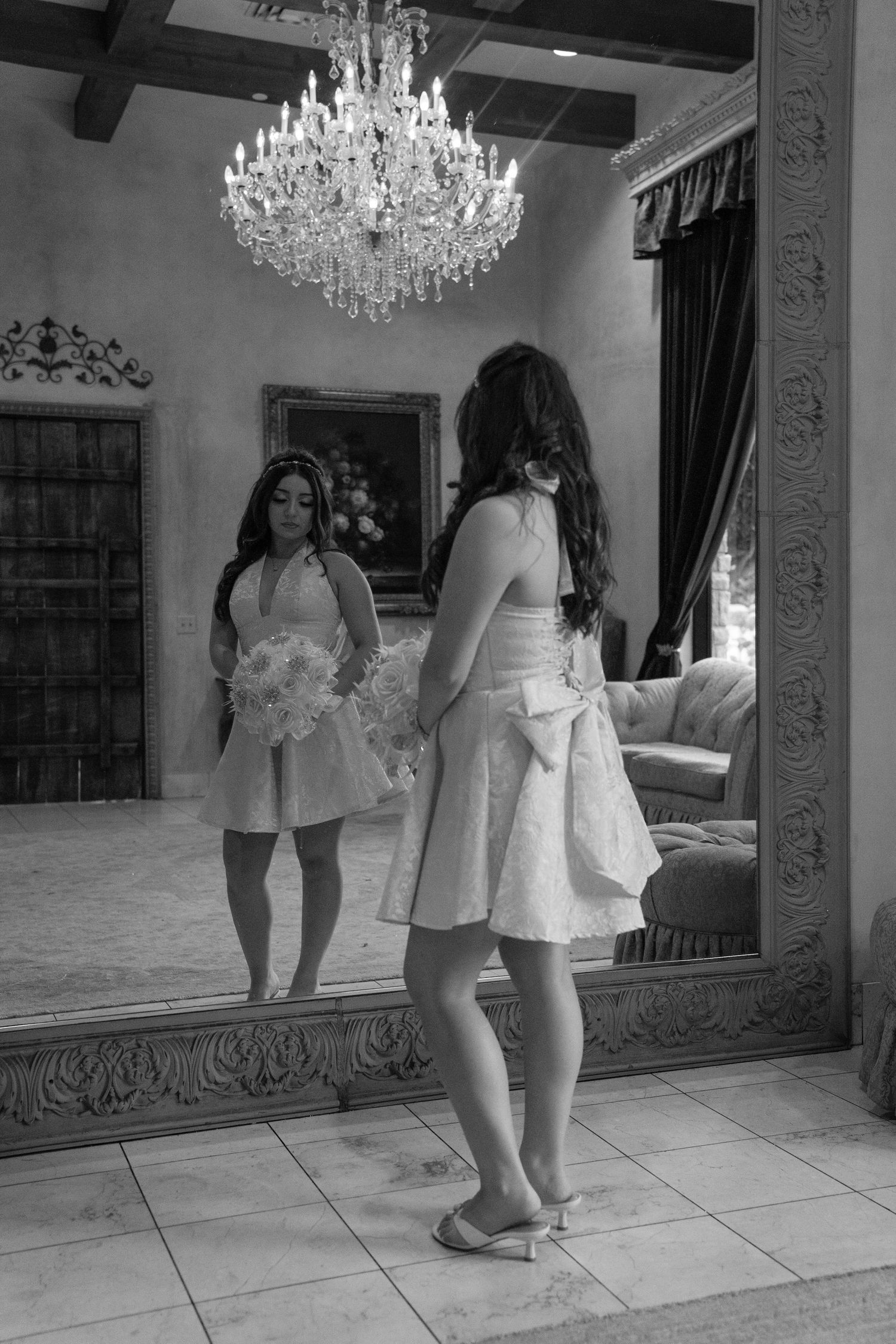 Black and white photo of woman in pink dress holding bouquet reflected in ornate mirror.