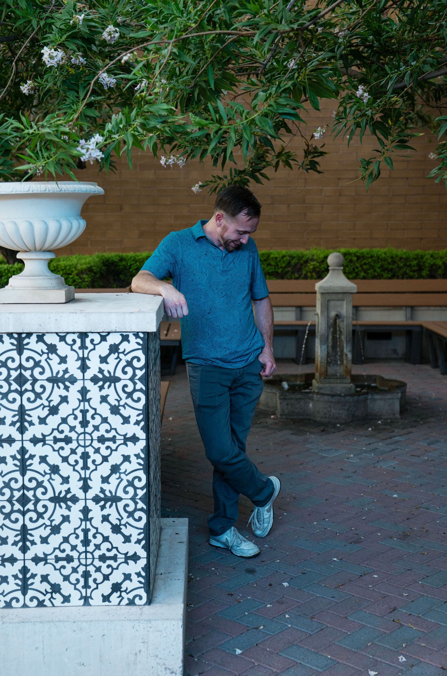 Man in teal polo leaning on ornate tiled wall in a garden courtyard with fountain.