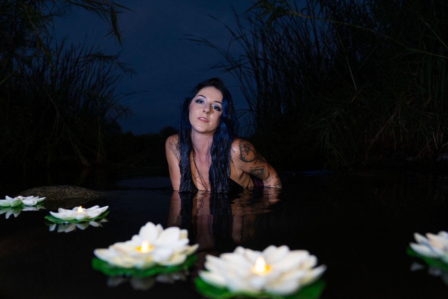 Tattooed woman emerging from dark water surrounded by floating white water lily flowers at night.