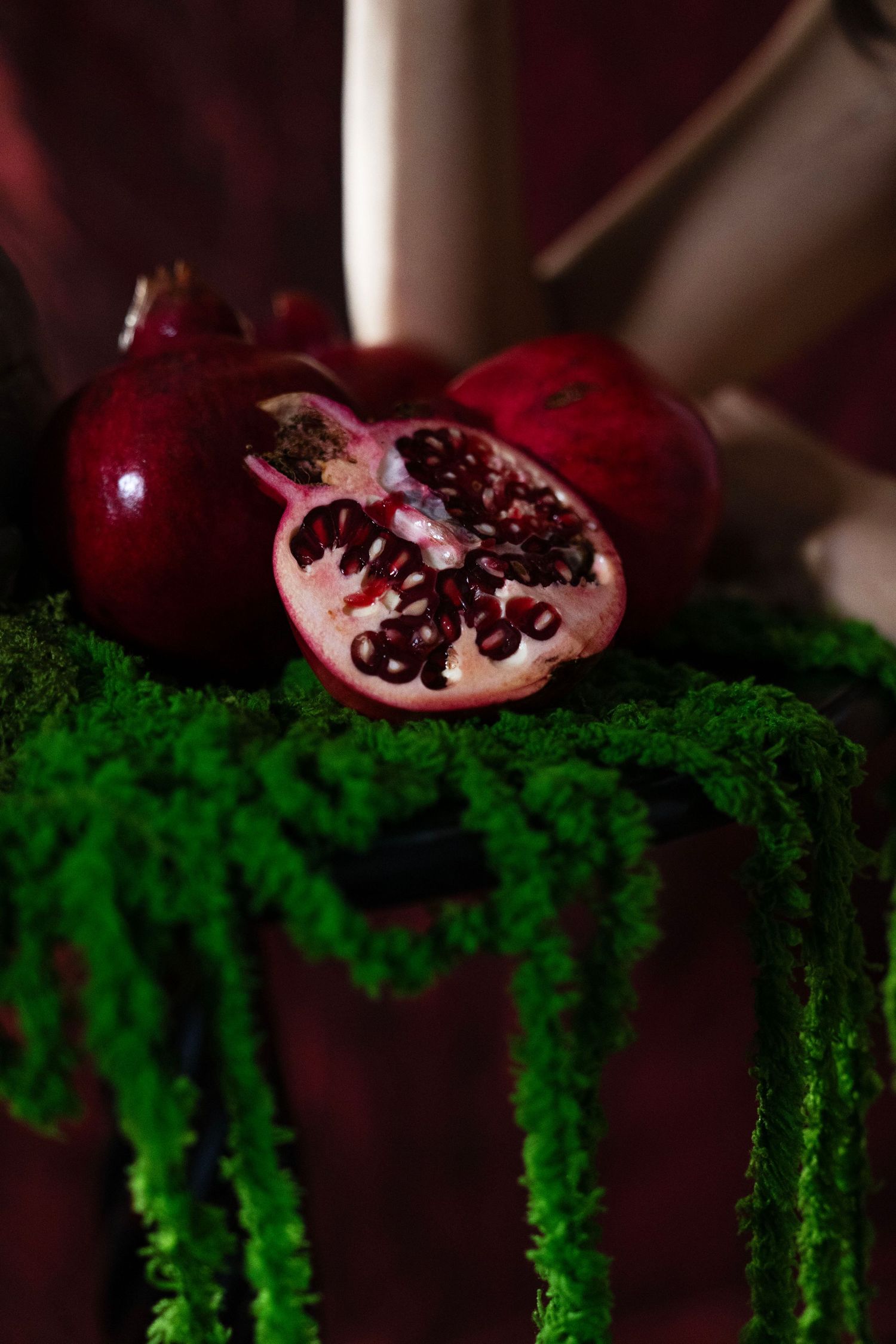 Halved ripe pomegranate with ruby red seeds beside whole pomegranates on a velvet table draped with bright green moss.