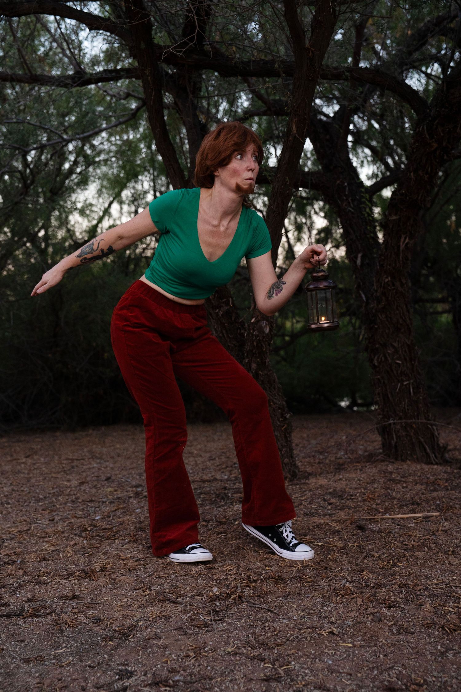 Woman in a green crop top, red velvet flared pants and black sneakers crouches in a forest at dusk, holding a glowing lantern.