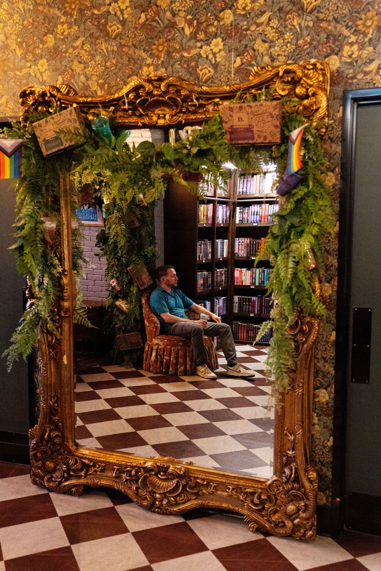 Man sitting in chair reflected in ornate gold mirror in cozy bookstore with lush green plants.