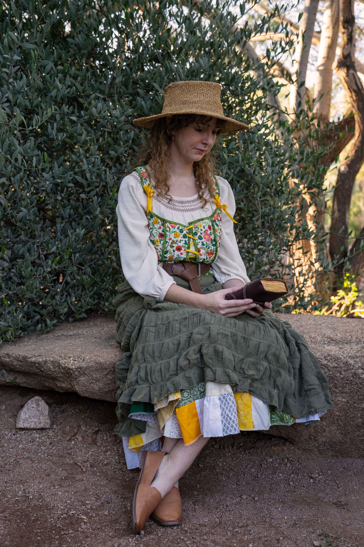 Woman in a straw hat and green ruffled skirt with a floral bodice sits on a stone bench outdoors, reading a book beside leafy bushes.