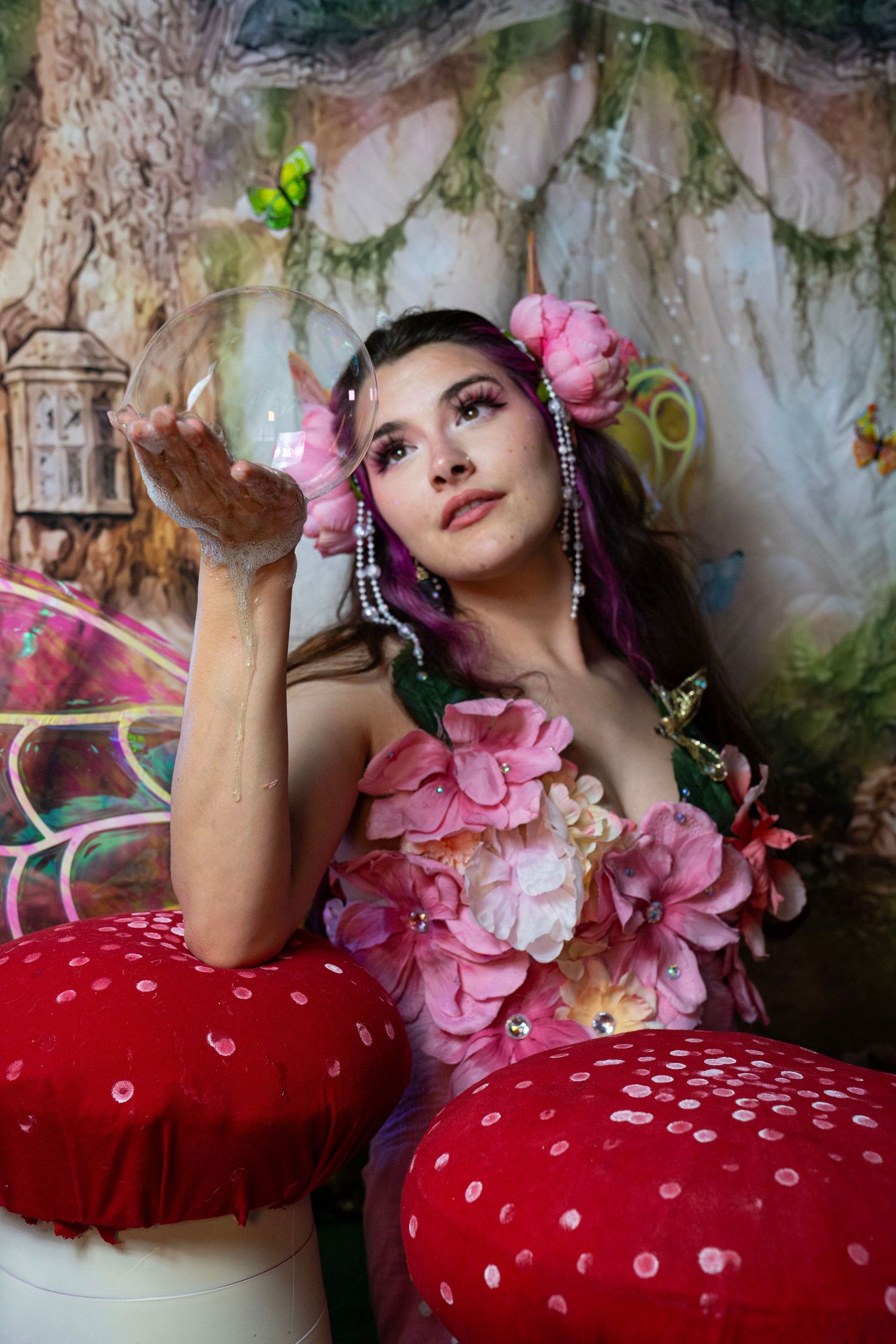 Fairy cosplay model adorned with pink flowers and iridescent wings poses between giant red mushrooms in fantasy set. Fantasy Photoshoot