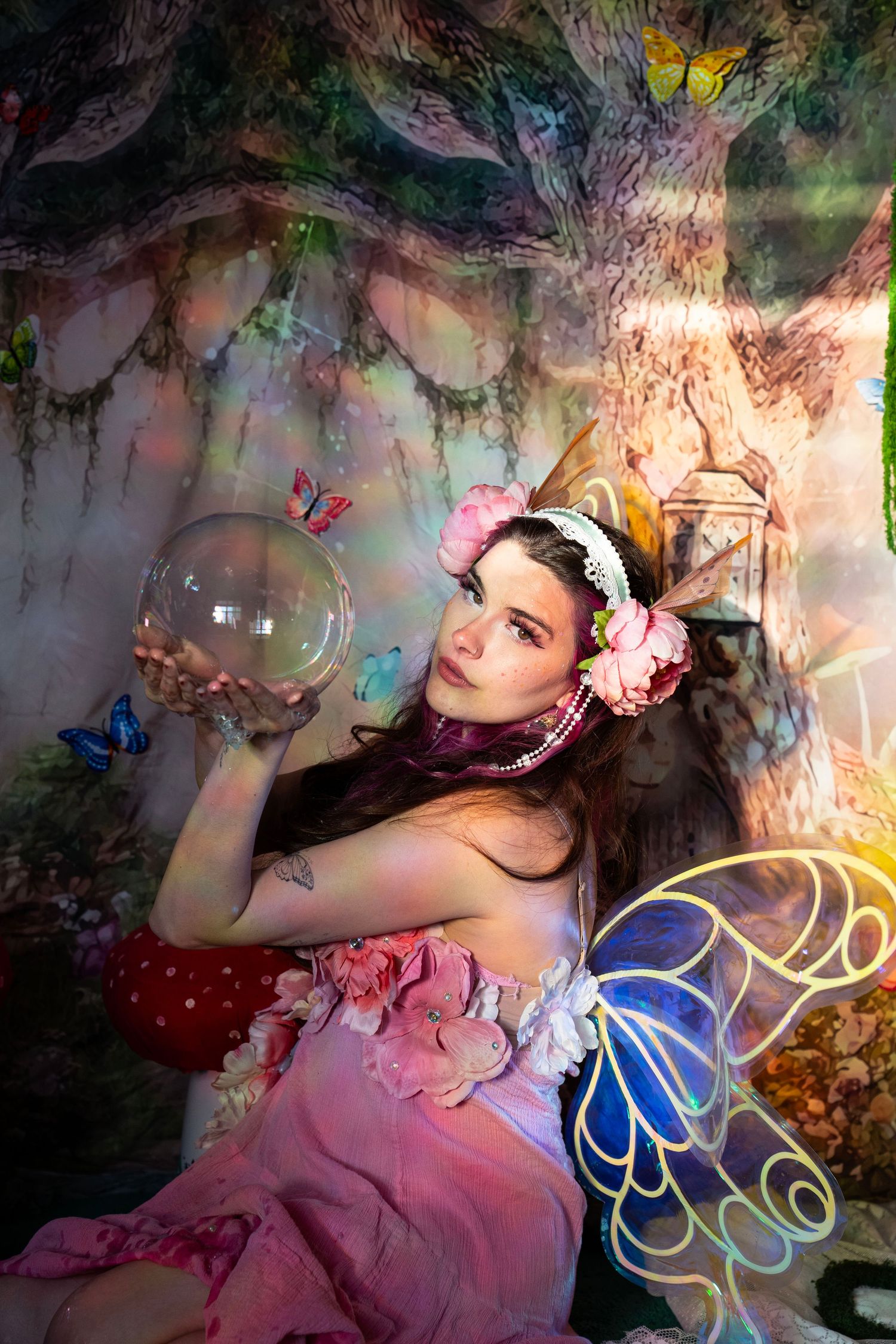 Woman in pink fairy dress holds a glass orb while posing in a colorful enchanted forest studio set. Fantasy Photoshoot