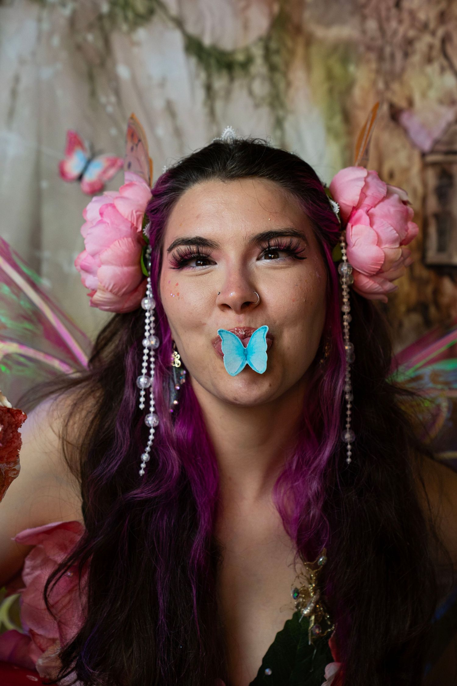 Woman in fairy costume with pink peony hair accessories holds blue butterfly prop in mouth, smiling playfully. Fantasy Photoshoot