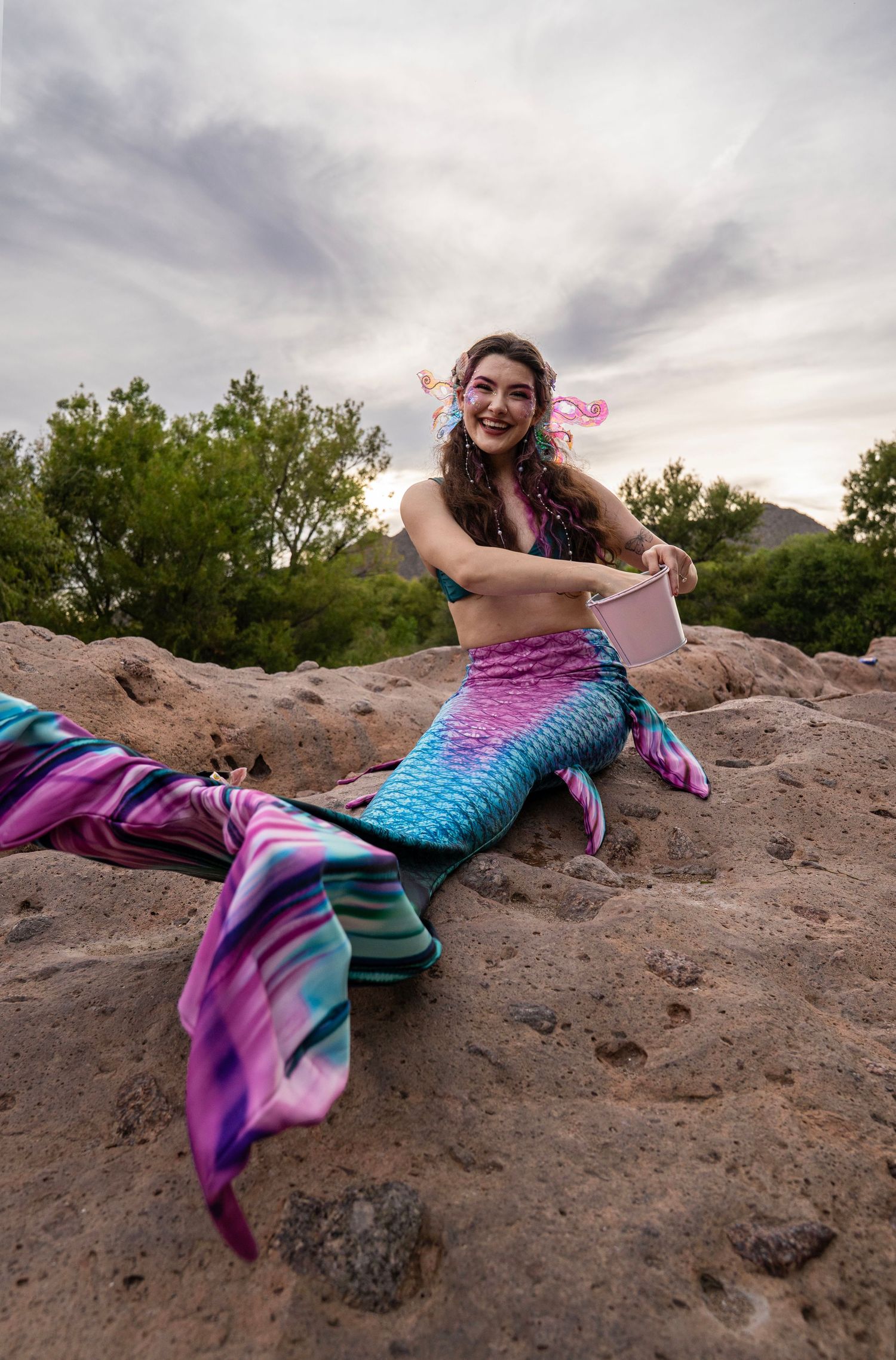 Smiling woman in a colorful mermaid costume sitting on rocks holding a small book under a cloudy sky. Fantasy Photoshoot