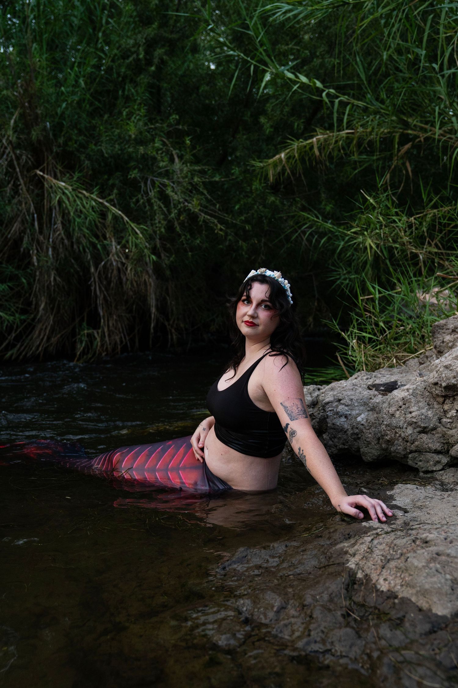 Dark-haired woman in black top with red mermaid tail and floral crown posing in river. Fantasy Photoshoot