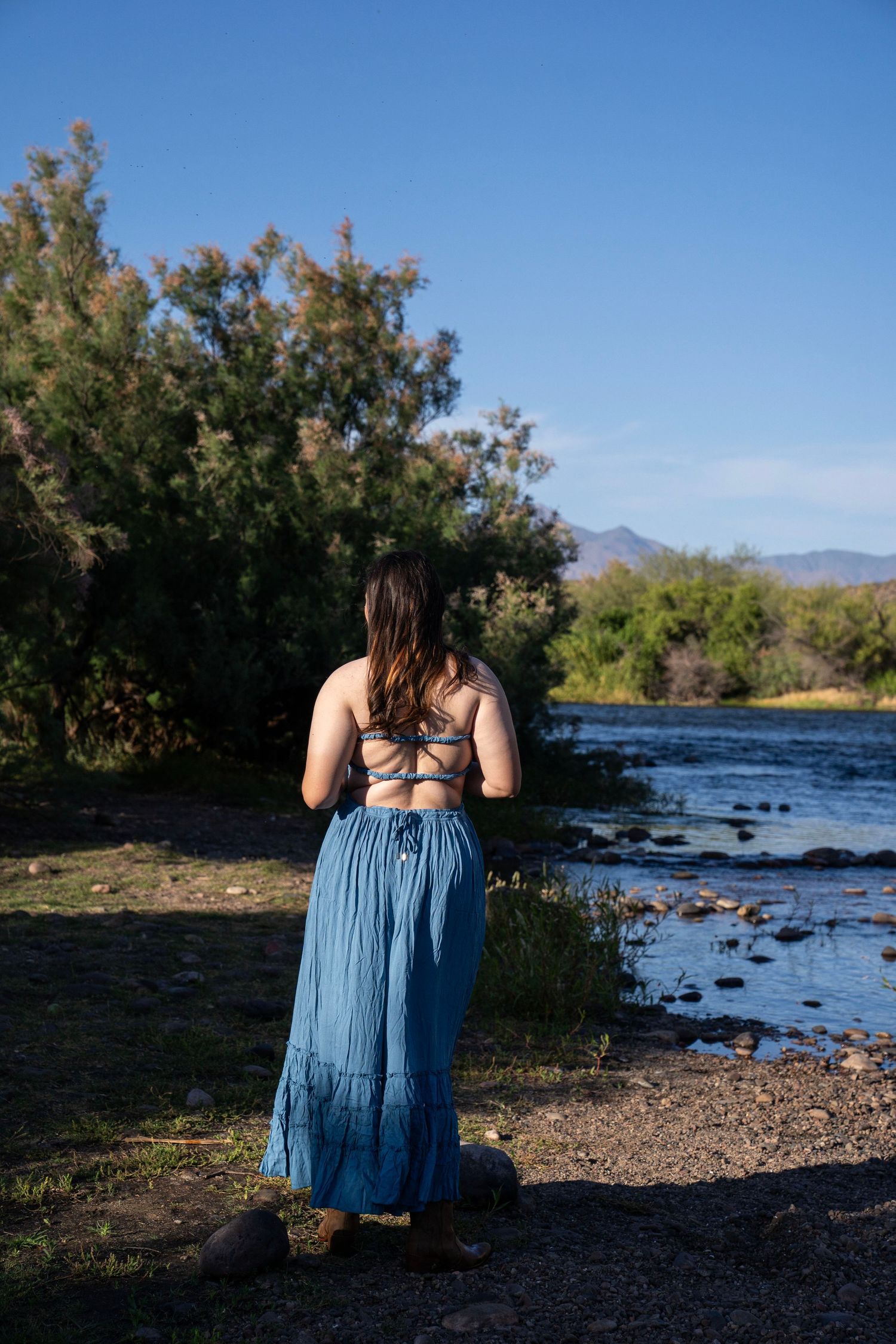 Woman in blue maxi skirt and crop top stands by a rocky riverbank surrounded by lush green trees and mountains.