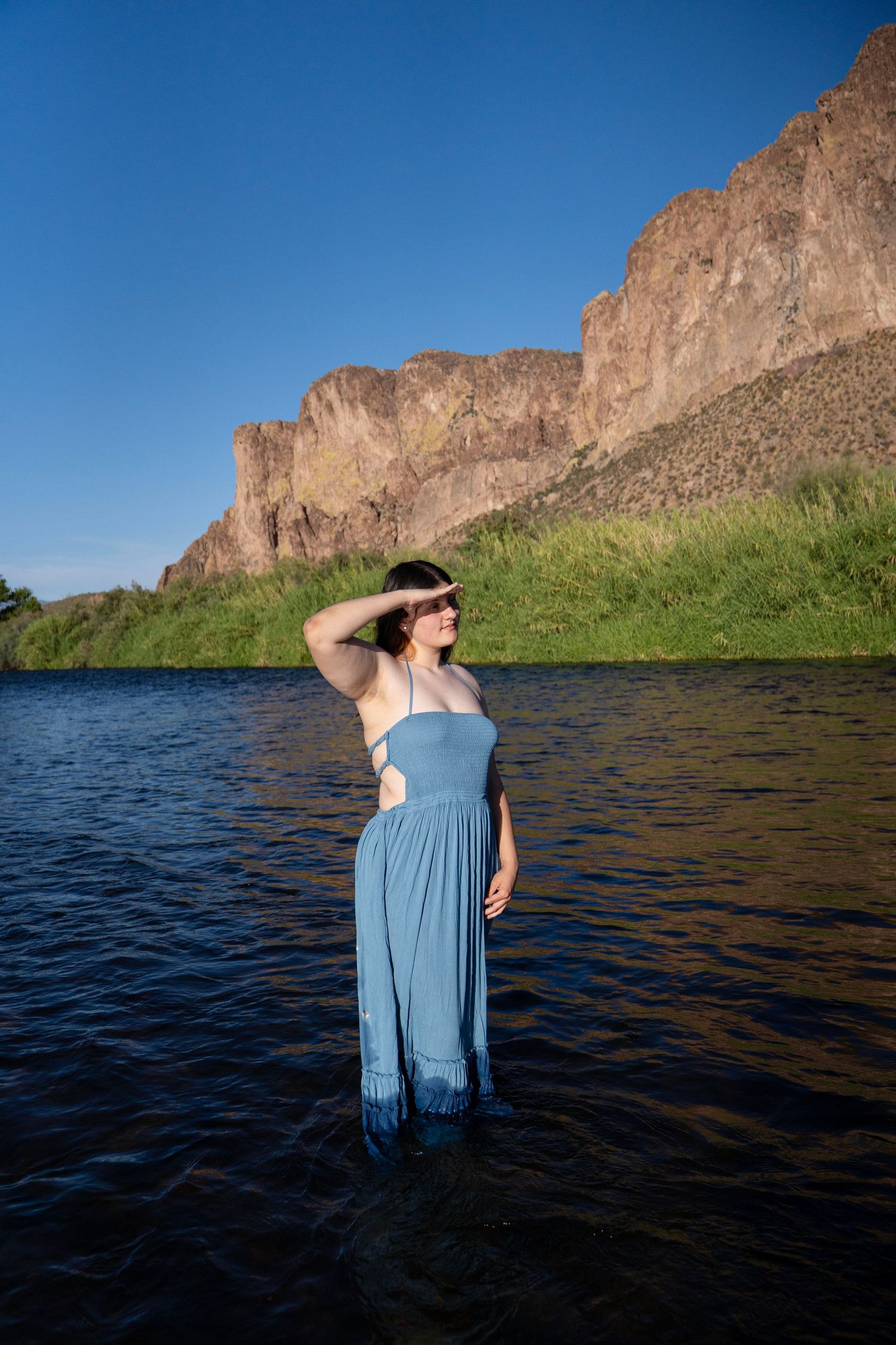 Woman in blue dress standing in river with rocky cliffs and green hills under clear blue sky.