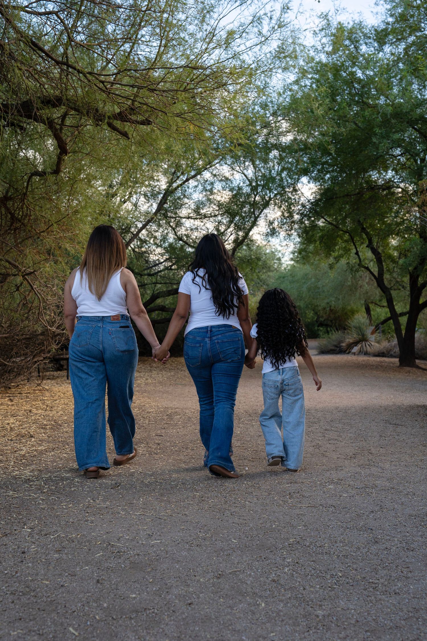 Three females in white shirts and jeans walk hand-in-hand down a tree-lined dirt path.