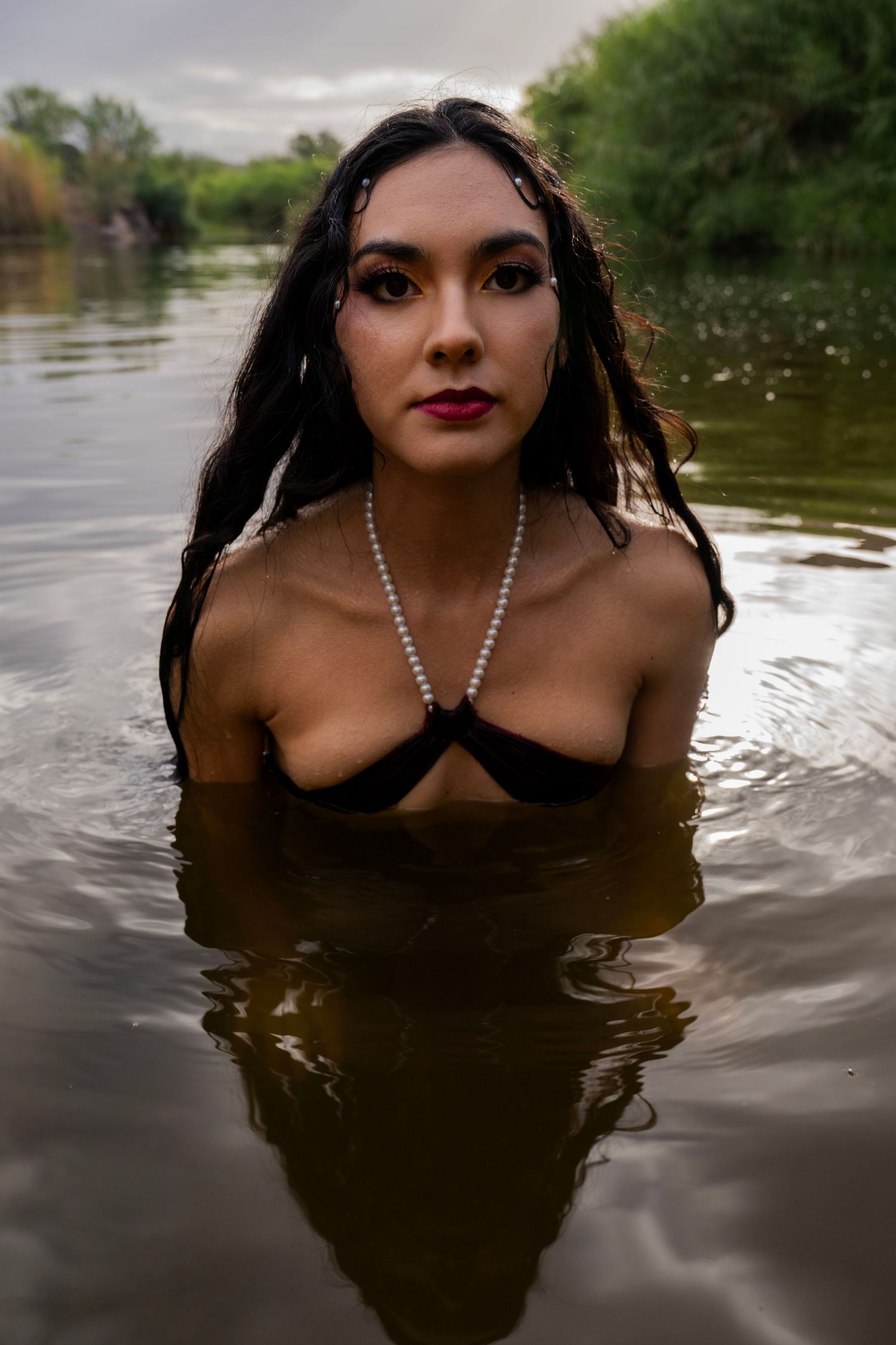 Dark-haired woman in black bikini and pearl necklace standing in a calm river with cloudy sky.