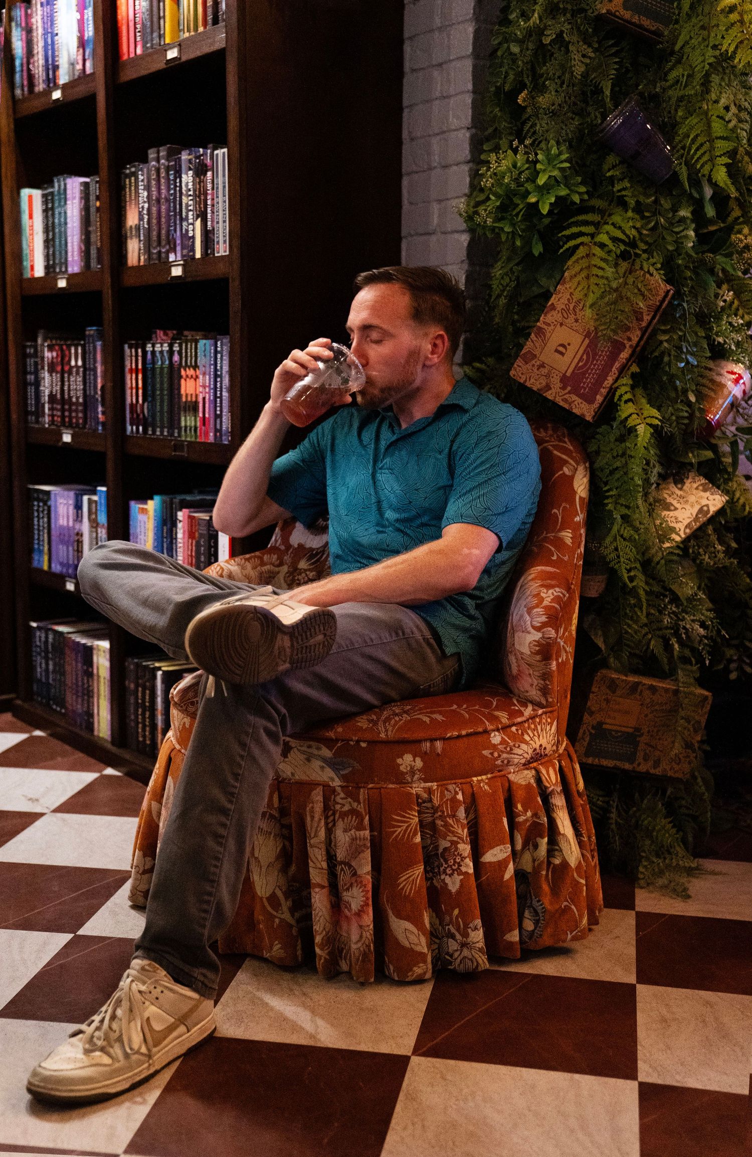 Man in teal polo sitting in floral armchair drinking water in a bookstore lounge.