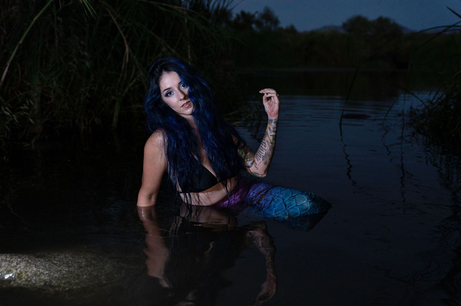Tattooed woman in mermaid costume sitting in dark water at night with colorful tail visible.
