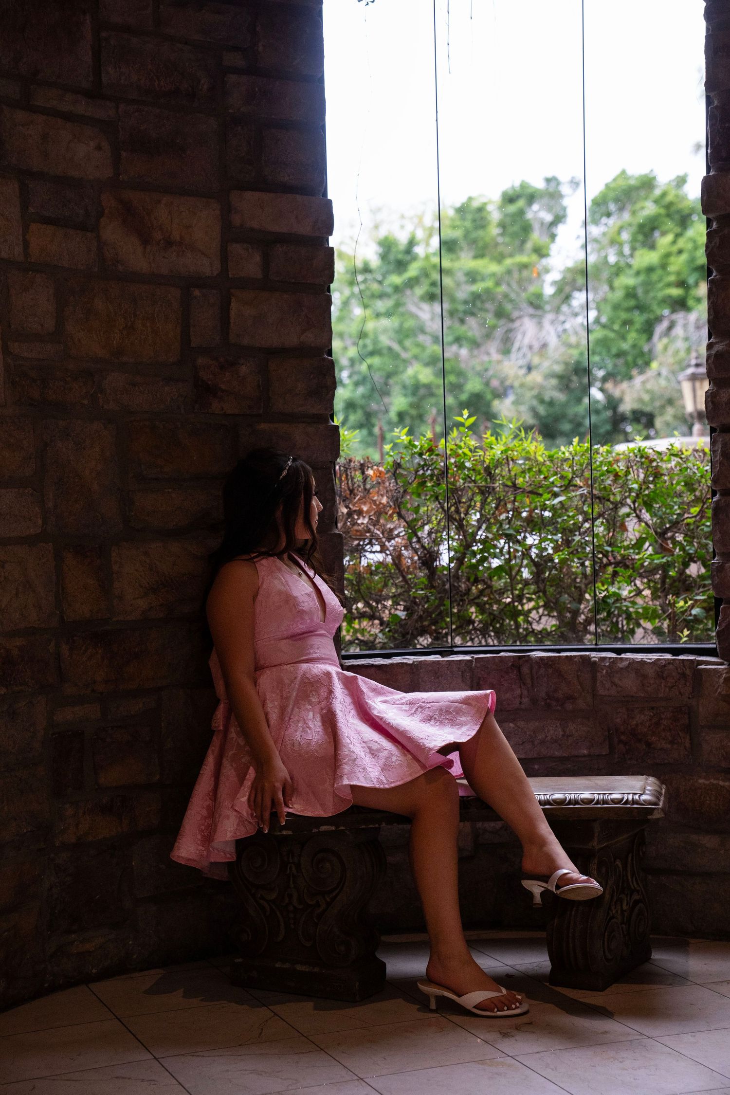 Young woman in pink dress leans against stone wall beside large window in historic building.